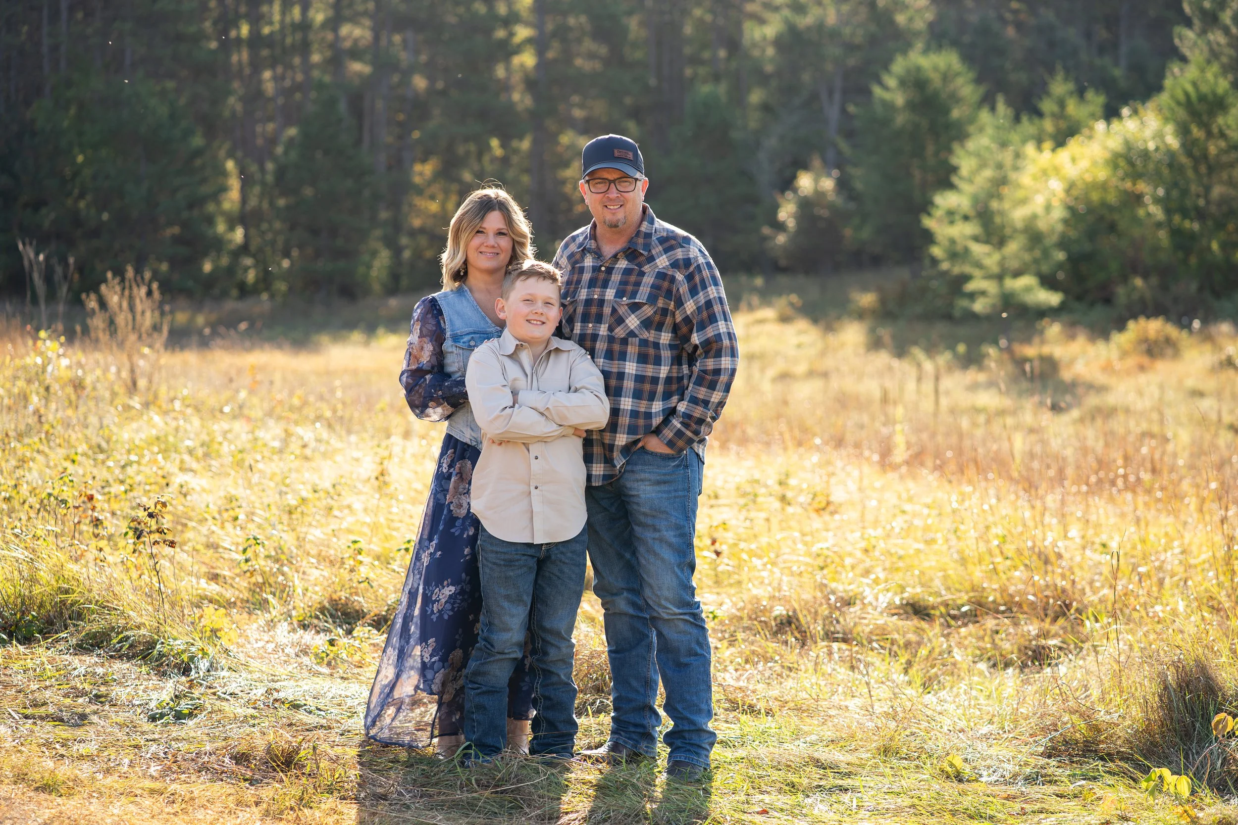 A family of three standing together outdoors in a scenic, grassy area during autumn. They are smiling and looking at the camera, with trees in the background.