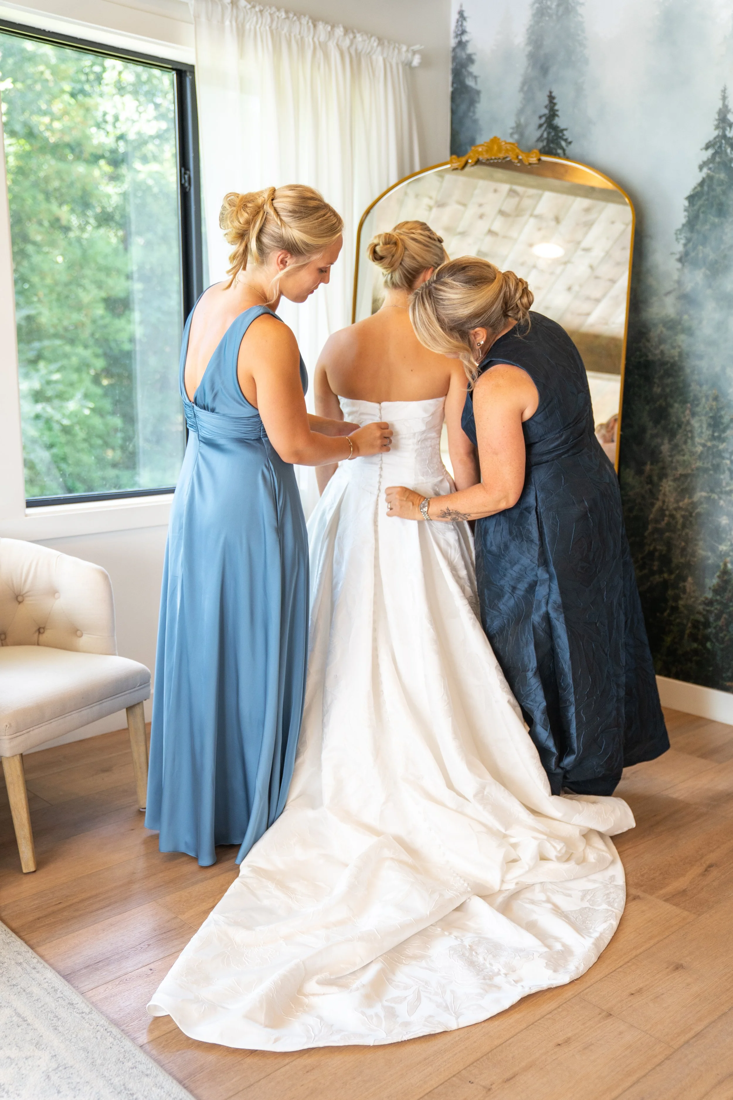 Three women, two in blue dresses and one in a black dress, helping a bride in a white wedding gown with her dress in a bright room with large windows, white curtains, and a mountain scene mural on the wall.