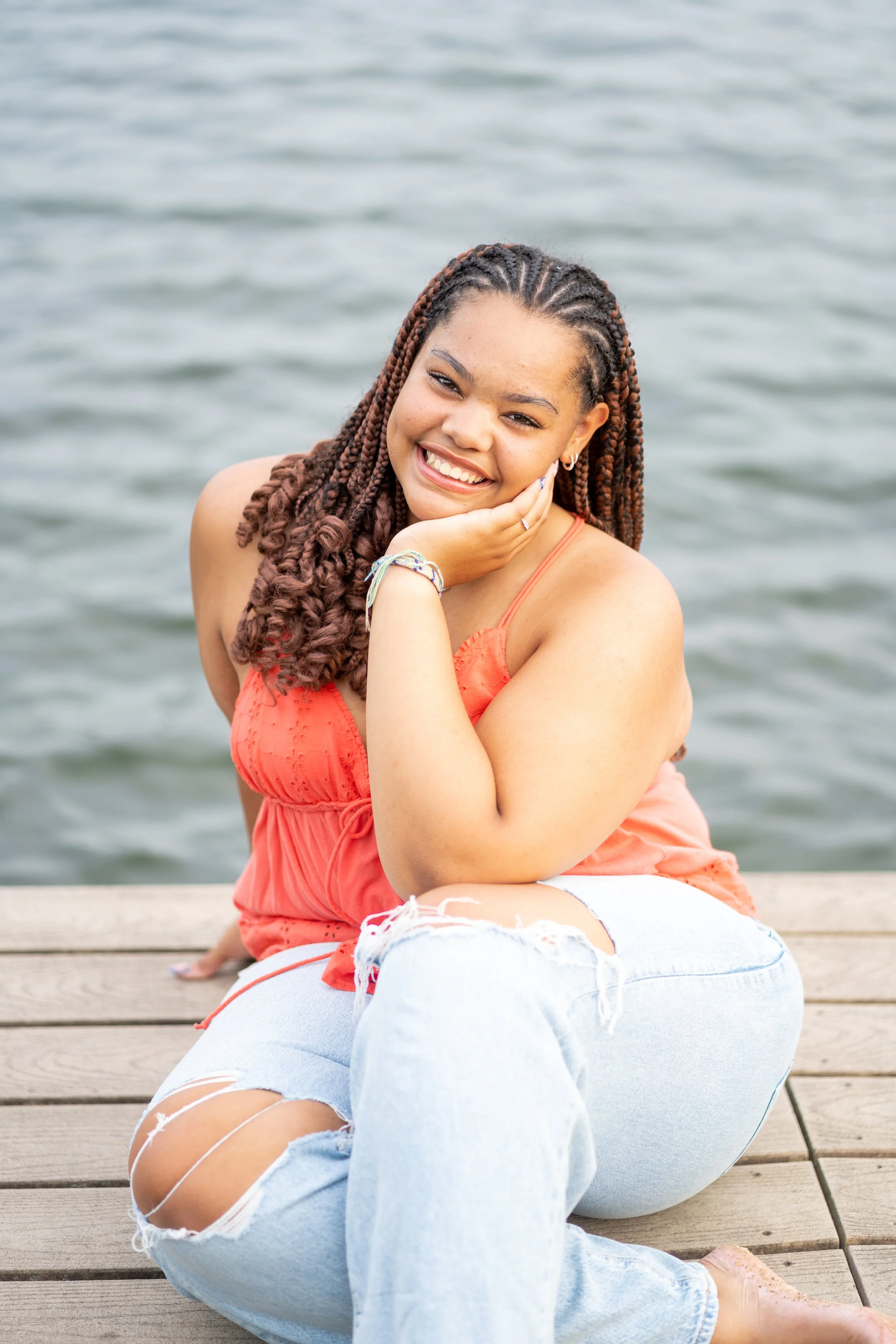A young woman with braided hair is sitting on a wooden dock near water, smiling, wearing an orange sleeveless top and ripped blue jeans.