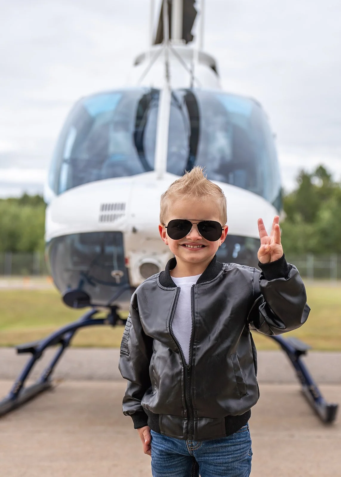 Child wearing sunglasses and a leather jacket making a peace sign in front of a helicopter.