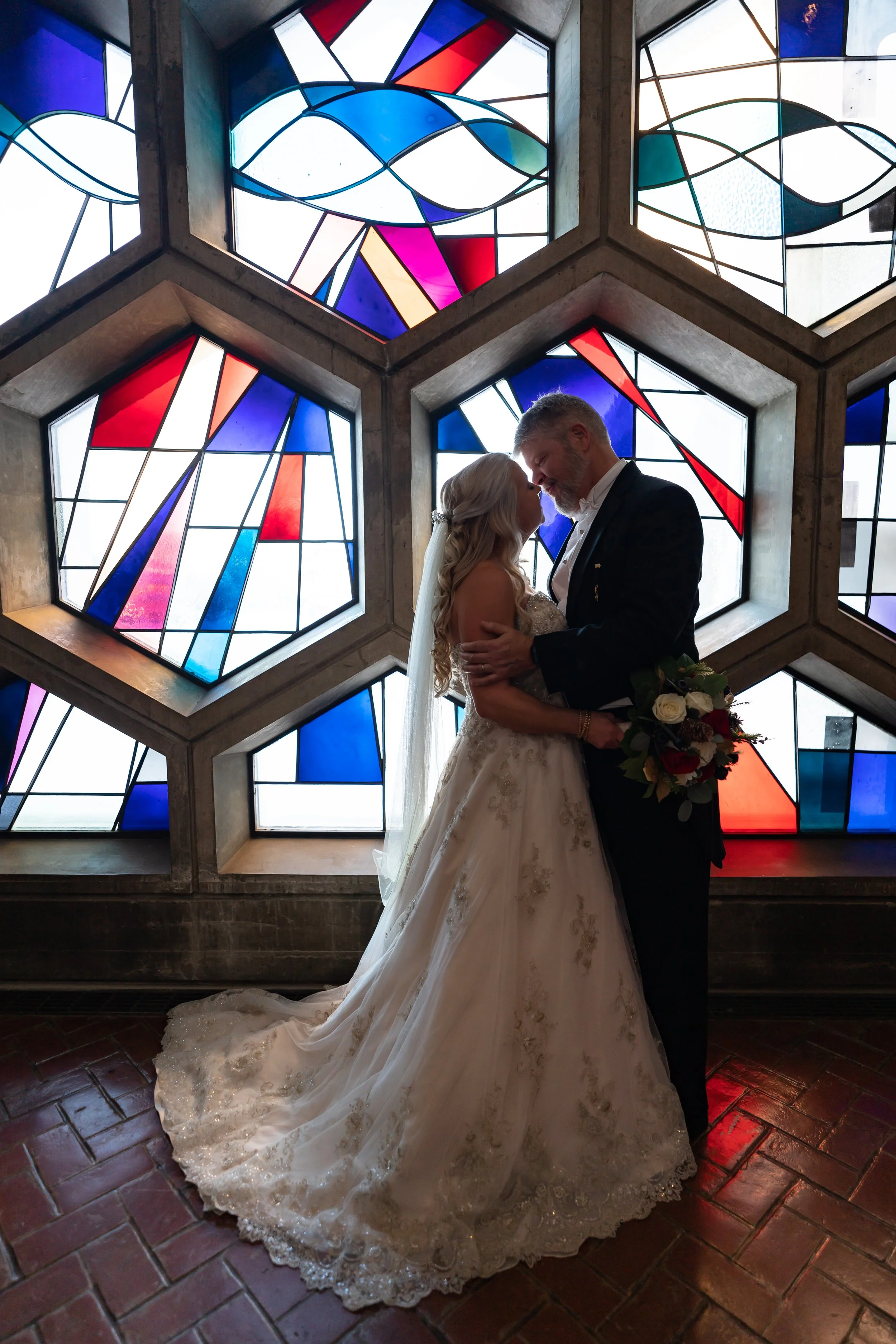 A bride and groom share a kiss in front of stained glass windows with colorful abstract patterns. The bride wears a white wedding gown with intricate lace details and a long train, and holds a bouquet of roses. The groom is dressed in a black tuxedo with a white shirt and bow tie. The setting appears to be indoors with a brick floor.