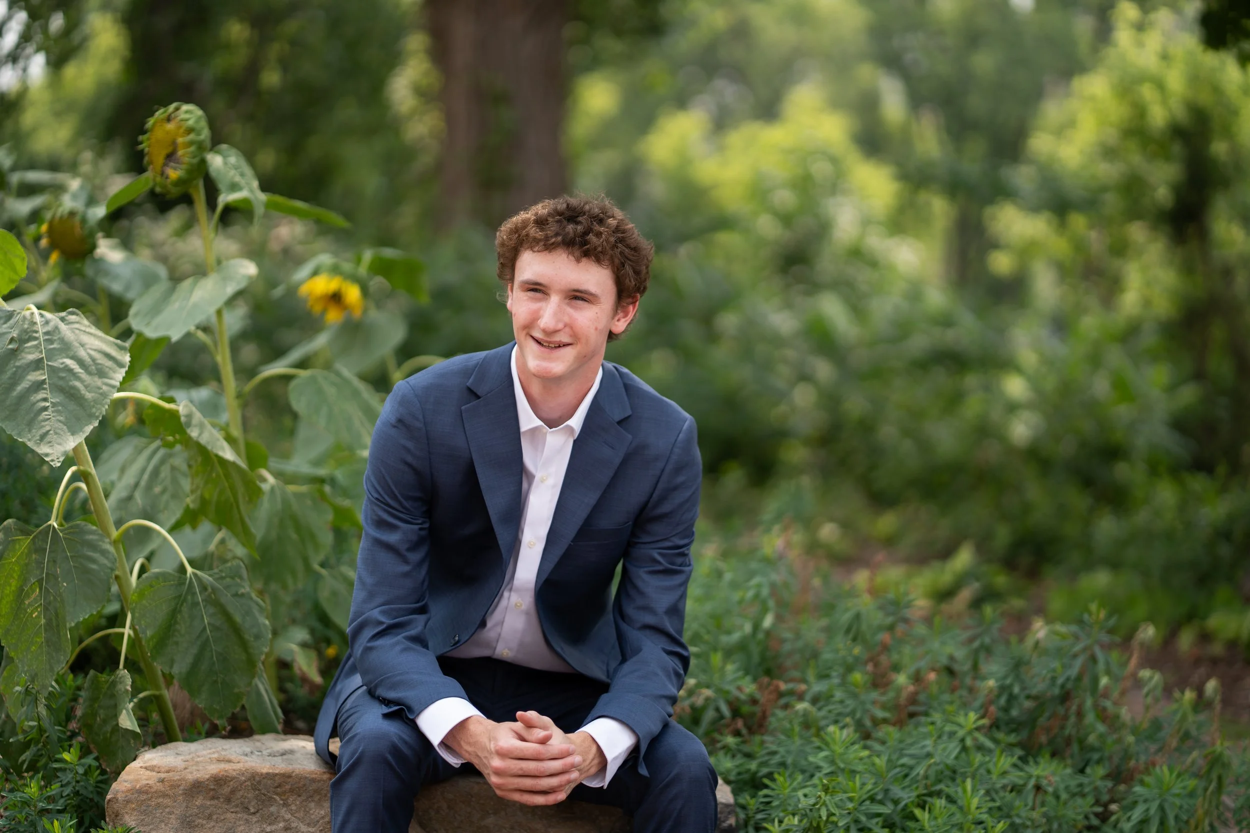 A young man in a blue suit sitting outdoors on a large rock surrounded by green plants and trees, smiling and looking to his right.