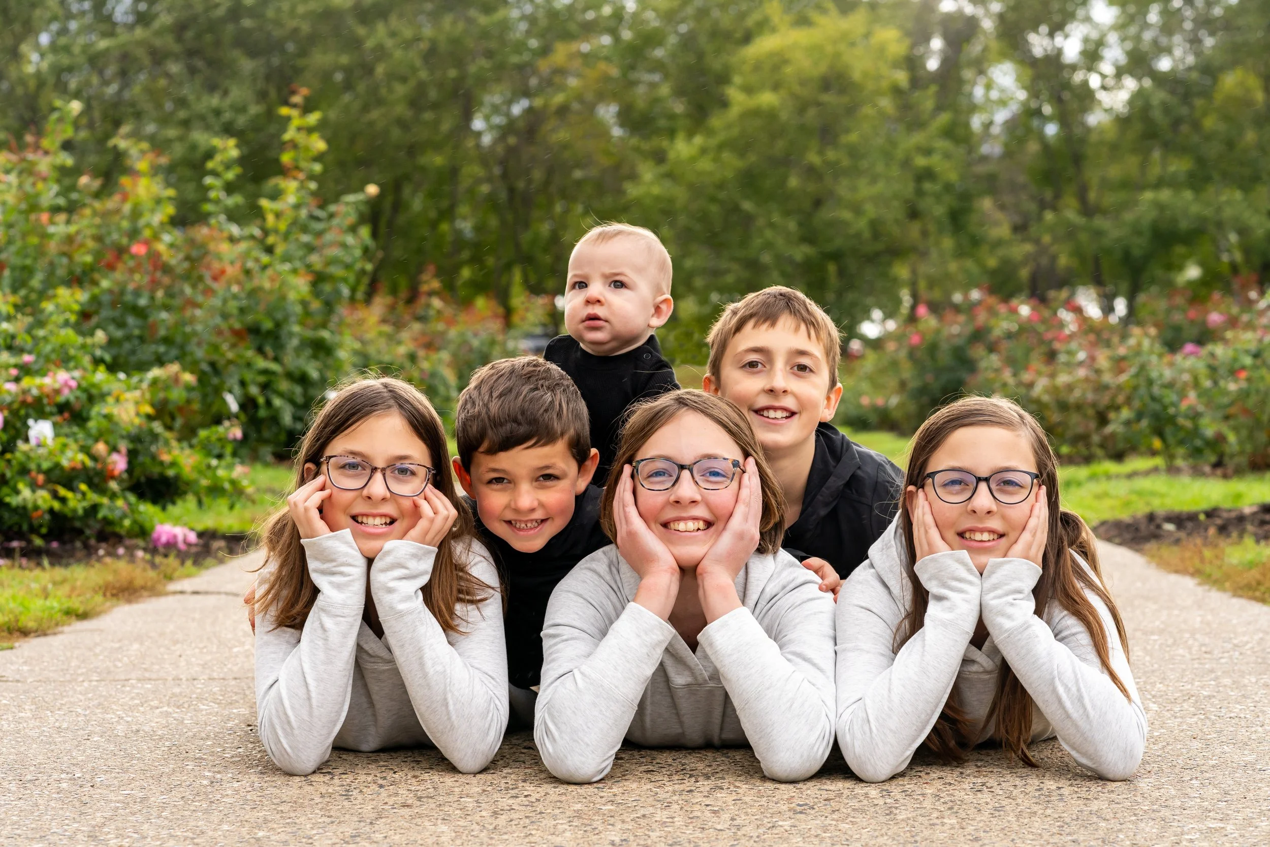 A group of six children, five girls and one boy, lying on the sidewalk in a park with trees and bushes in the background during daytime. The children are smiling and looking at the camera, with some resting their heads on their hands or cheeks.