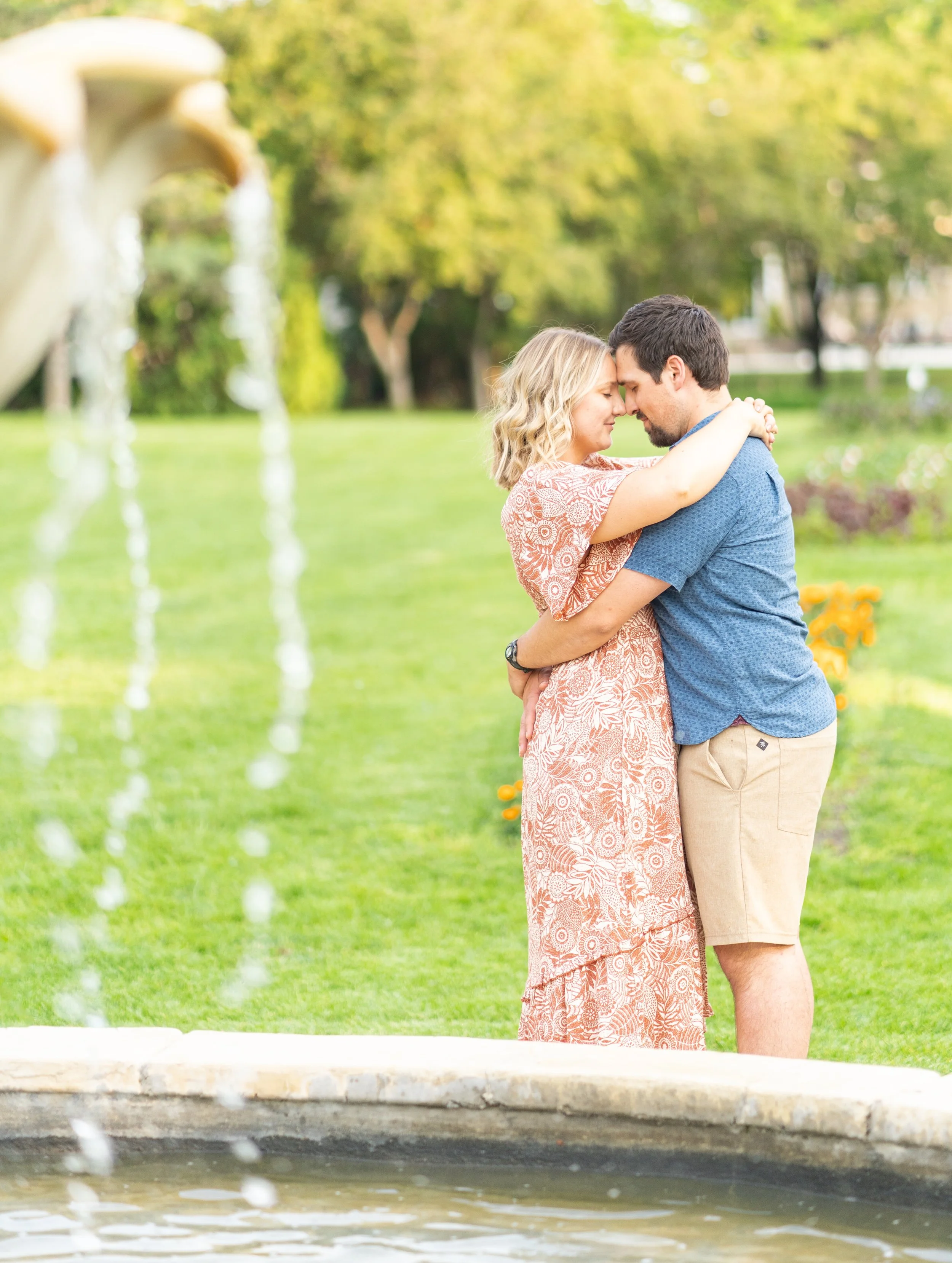 A couple embraces with foreheads touching, standing outdoors by a fountain with water streaming down, in a park with green grass and trees in the background.