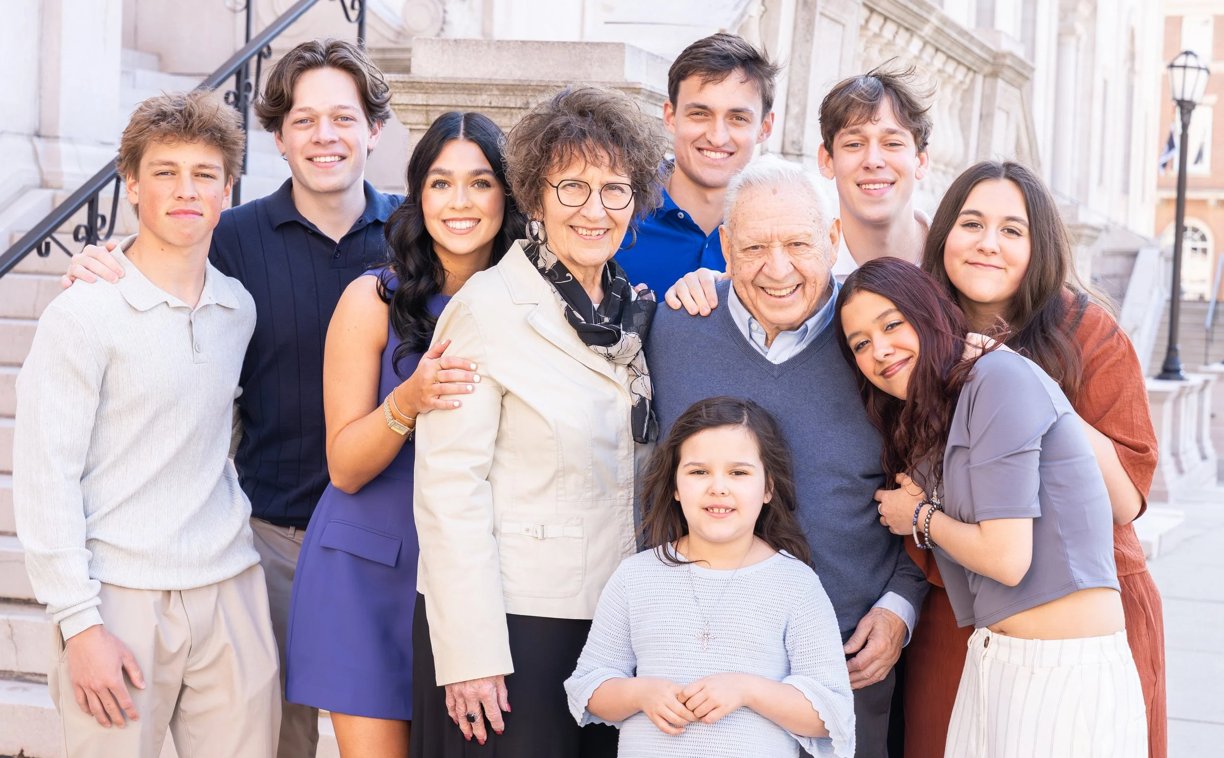 A diverse group of adults and children smiling and posing together outside on a staircase in front of a historic building.