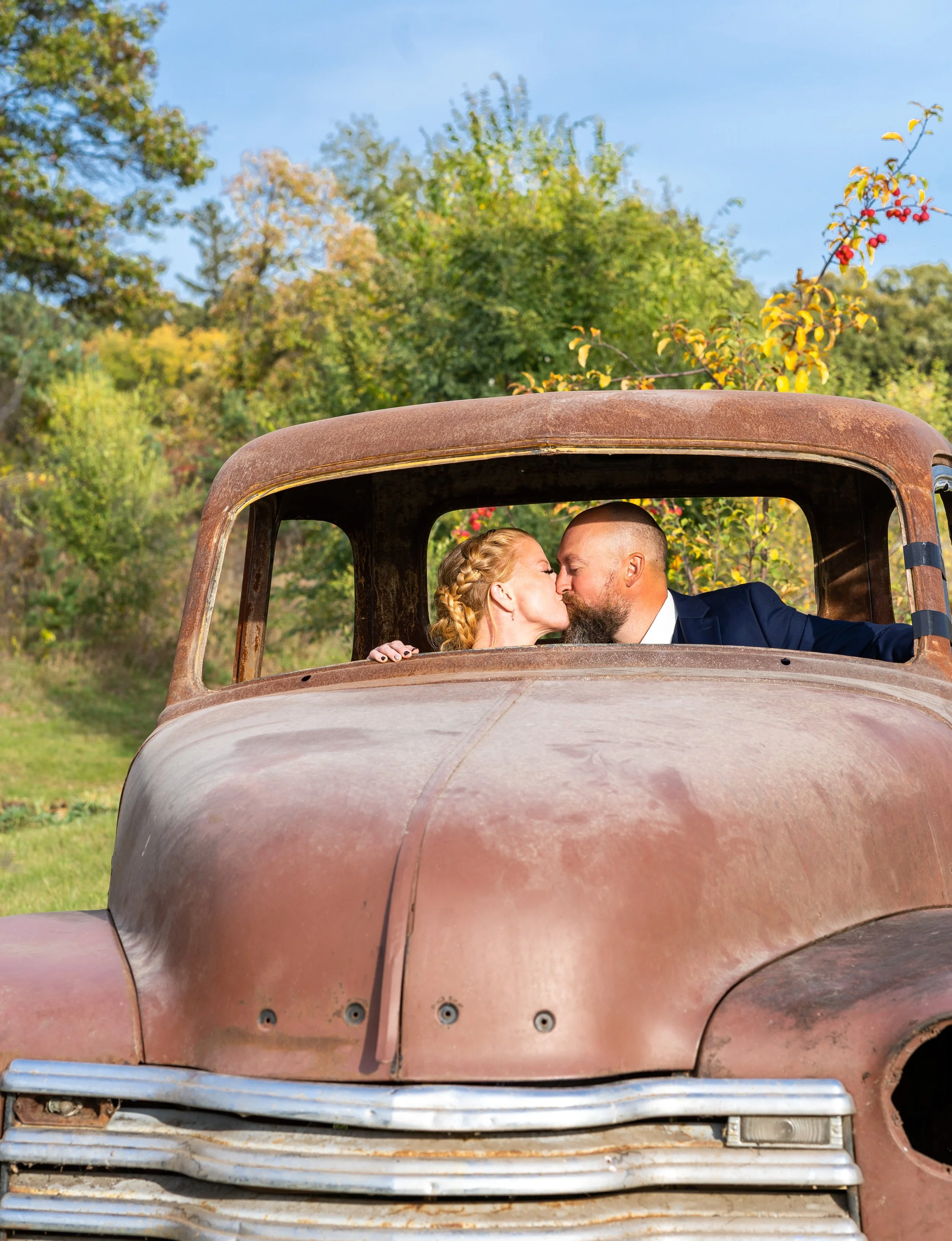 A couple in wedding attire, a woman with braided hair and a man with a shaved head, kissing inside an old, rusty truck with a rusted hood. The background features green trees and a blue sky.