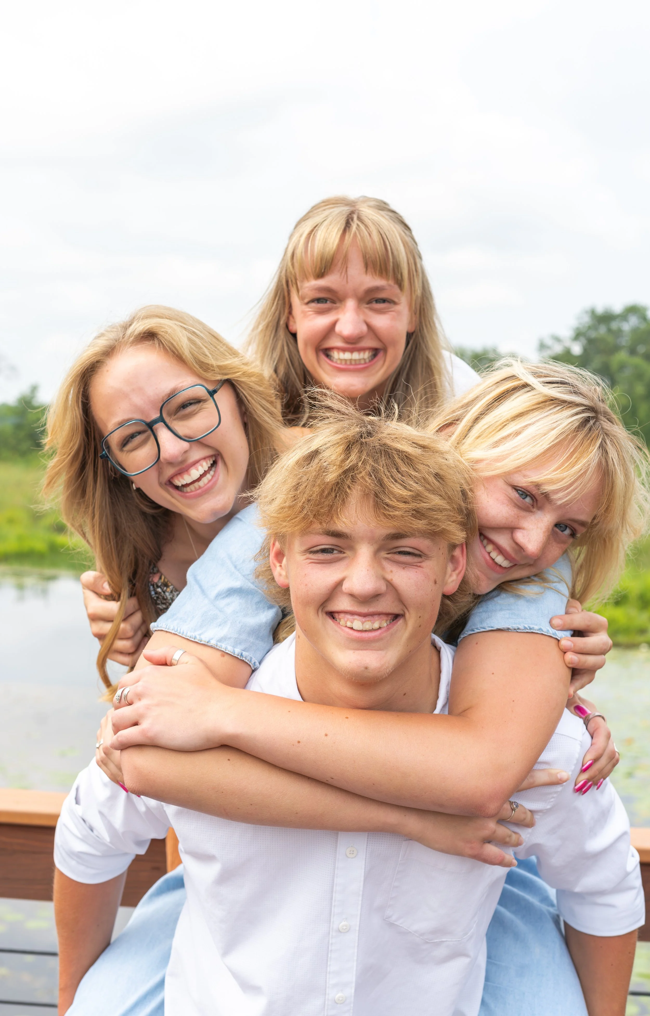 A group of five young adults smiling and hugging outdoors near a body of water with trees in the background.