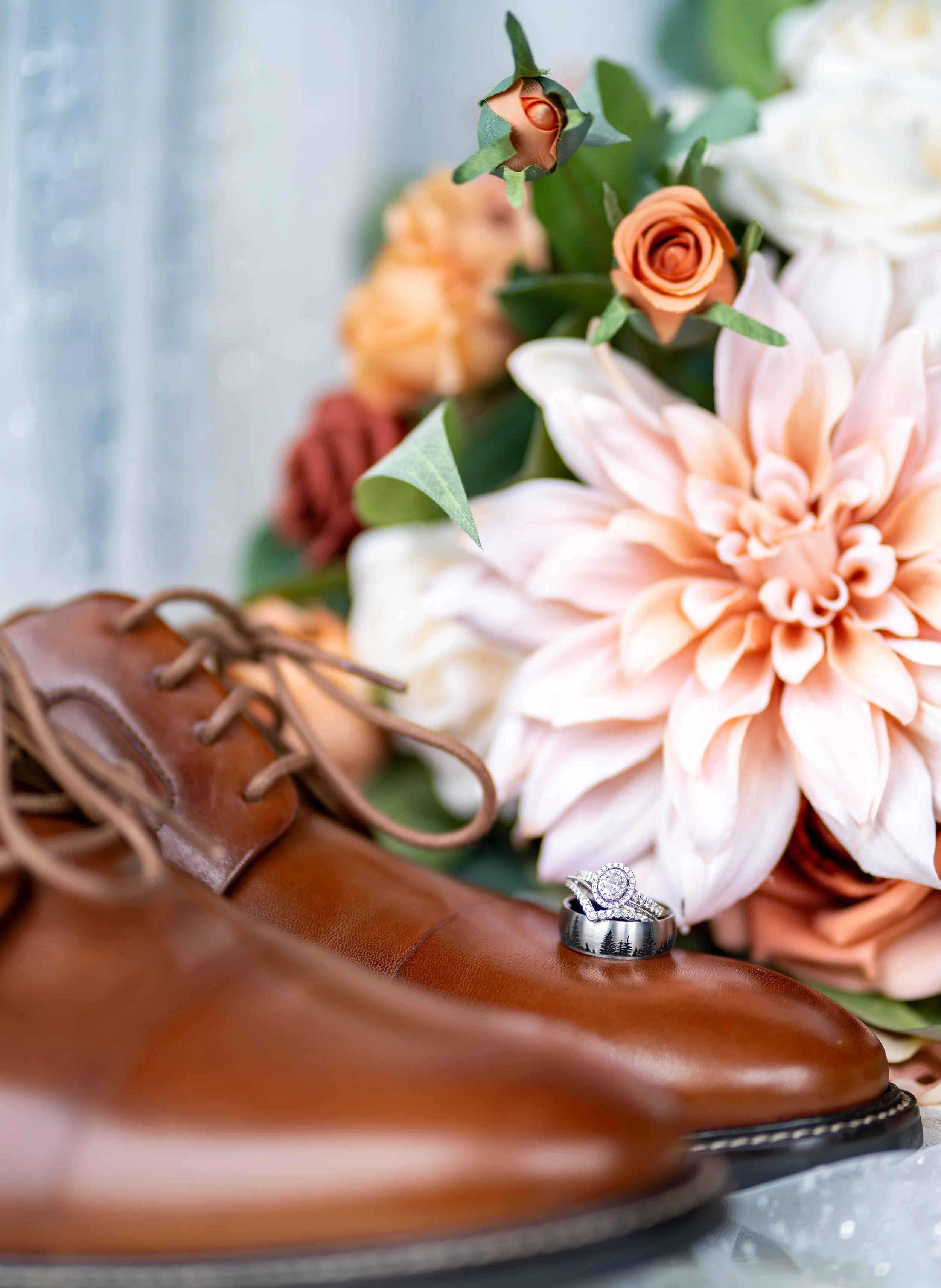 A pair of brown leather dress shoes, a bouquet of peach, white, and blush roses, and two engagement rings with diamonds, all arranged on a surface.