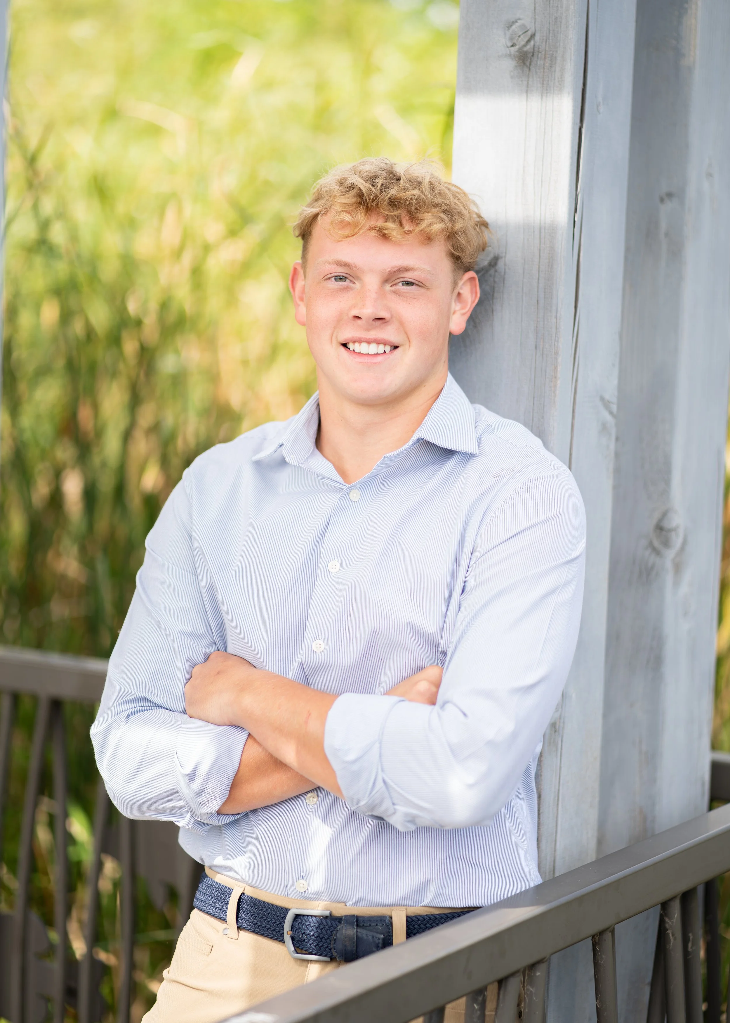 A young man with curly blonde hair, wearing a light blue, button-up shirt, beige pants, and a dark blue woven belt, standing outdoors with arms crossed, leaning against a gray wooden fence, with green foliage in the background.