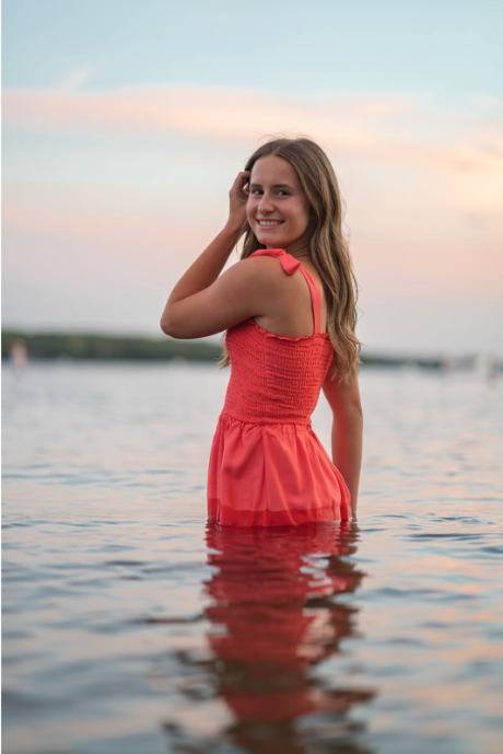 Young woman in a coral dress standing in calm water outdoors during sunset, smiling and looking at the camera.