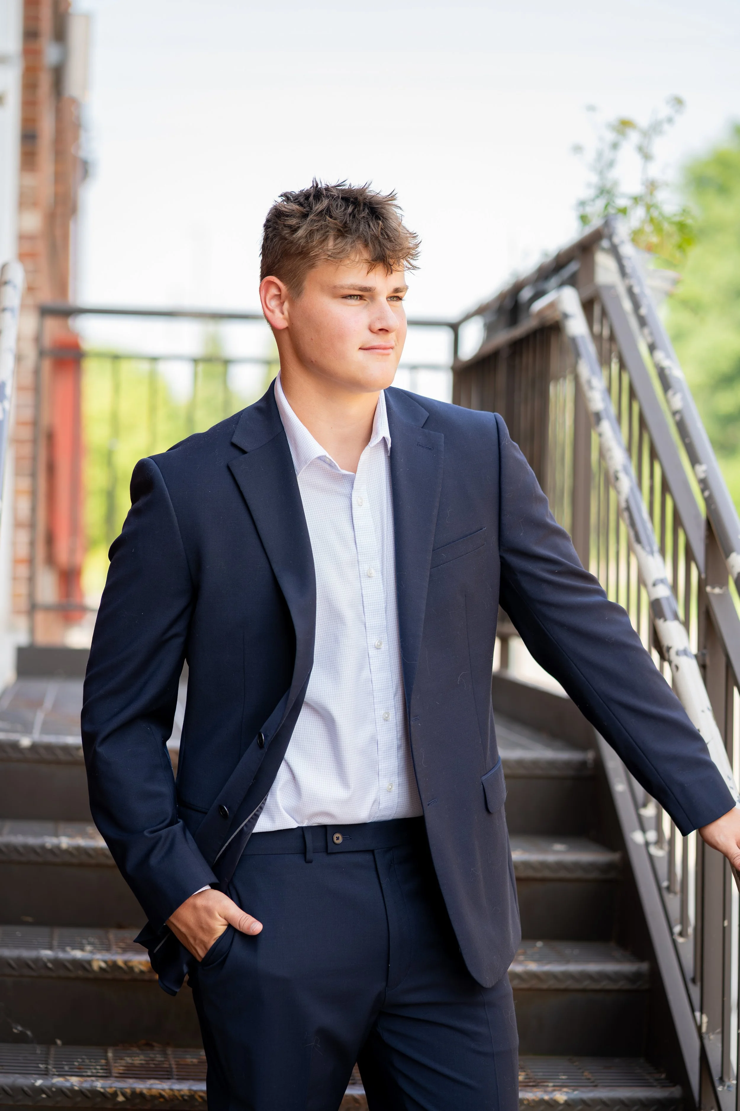 A young man in a navy suit and white shirt standing outdoors on a staircase railing with a relaxed pose and confident expression.