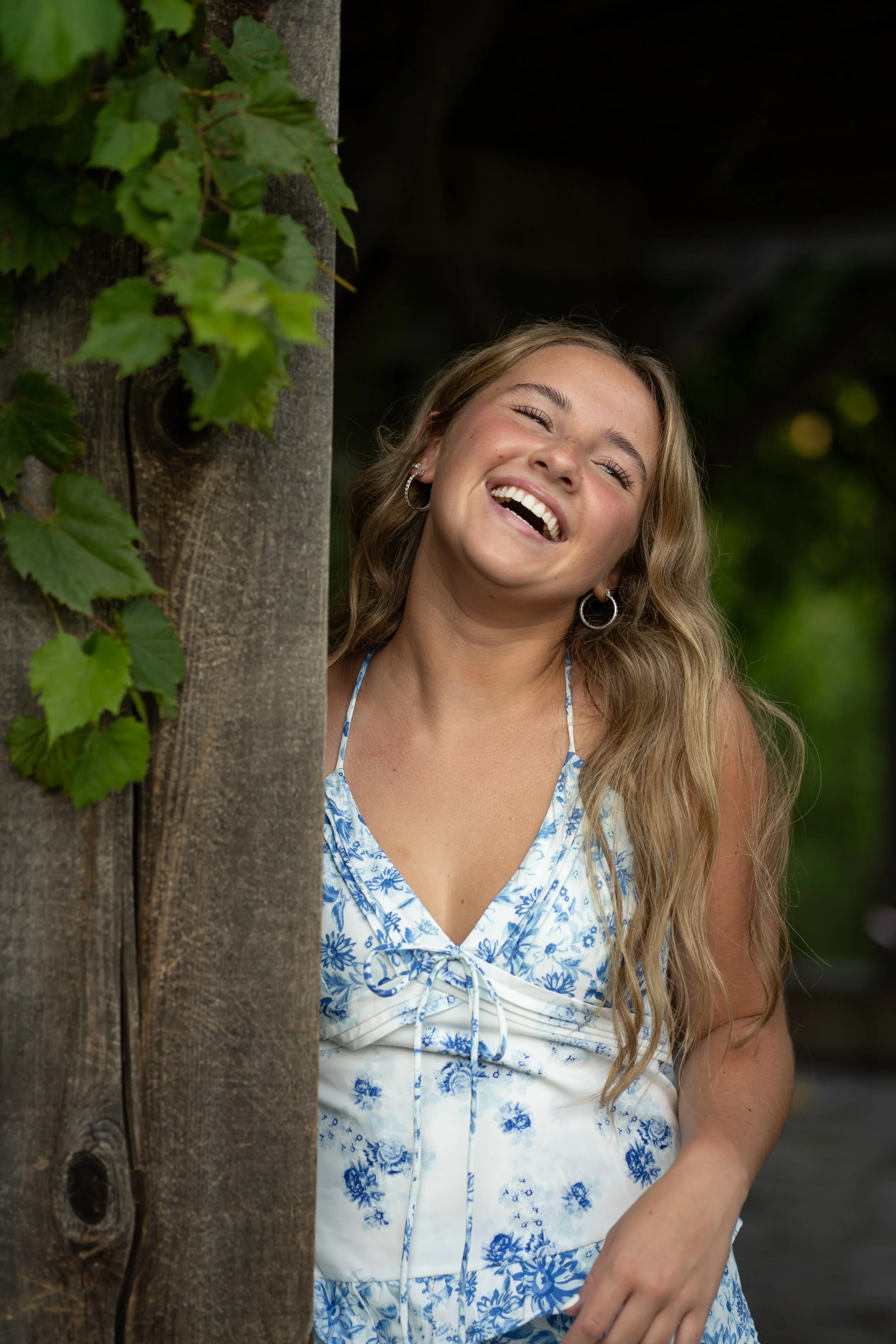 A young woman with long wavy blonde hair is smiling and laughing, standing next to a wooden post with green ivy leaves climbing on it, in an outdoor setting.