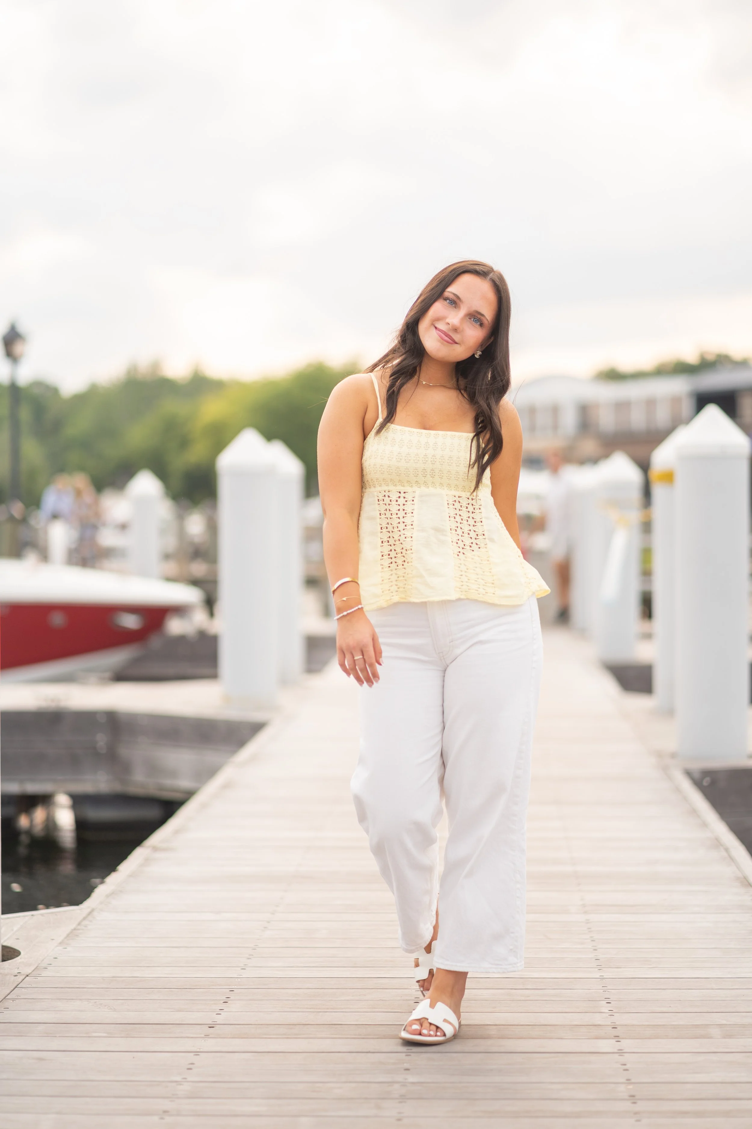 A woman with long dark hair wearing a cream-colored sleeveless top, white pants, and white sandals walking on a wooden dock near a marina with boats. The background has a cloudy sky and some greenery.