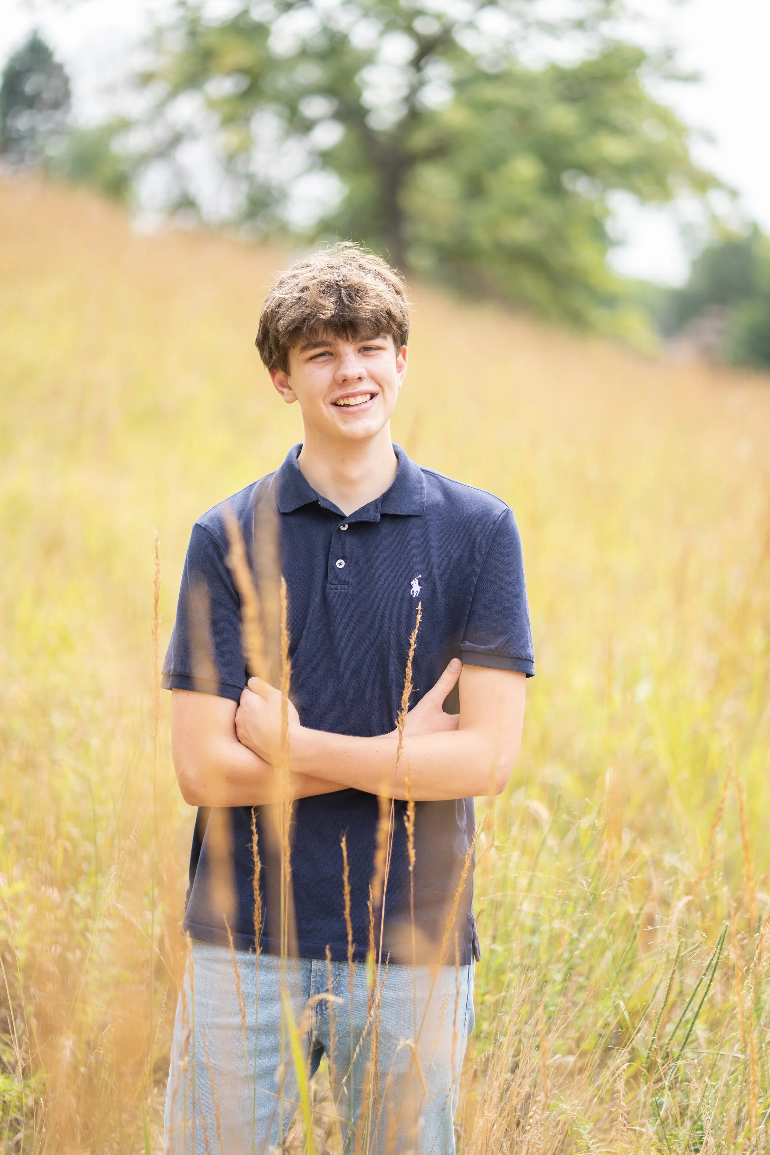 A young man with brown hair wearing a navy polo shirt and light-colored jeans standing in a field of tall, golden grass with trees in the background.