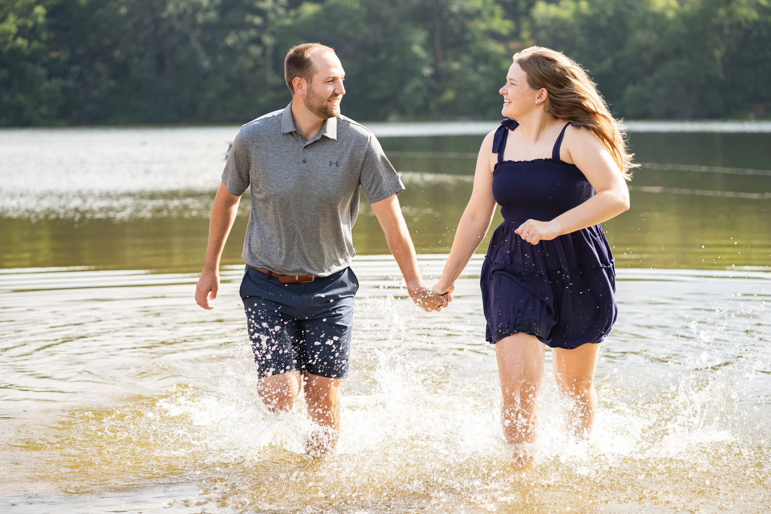 A man and woman holding hands and walking together in shallow water near a lake or river, smiling and looking at each other, with trees in the background.