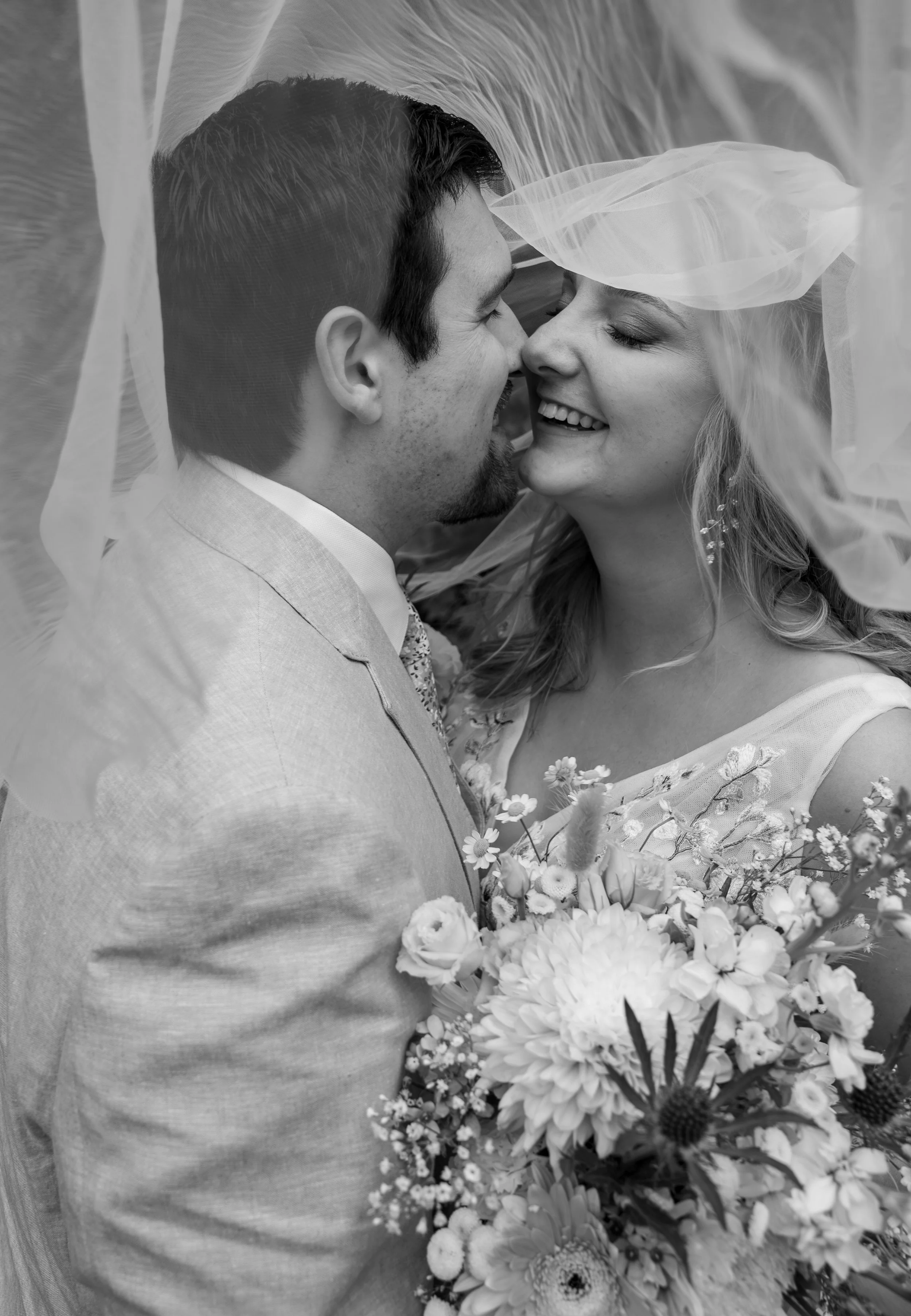 A black and white photograph of a bride and groom close together on their wedding day, smiling and about to kiss. The bride holds a large bouquet of flowers, and a veil drapes over both of them.