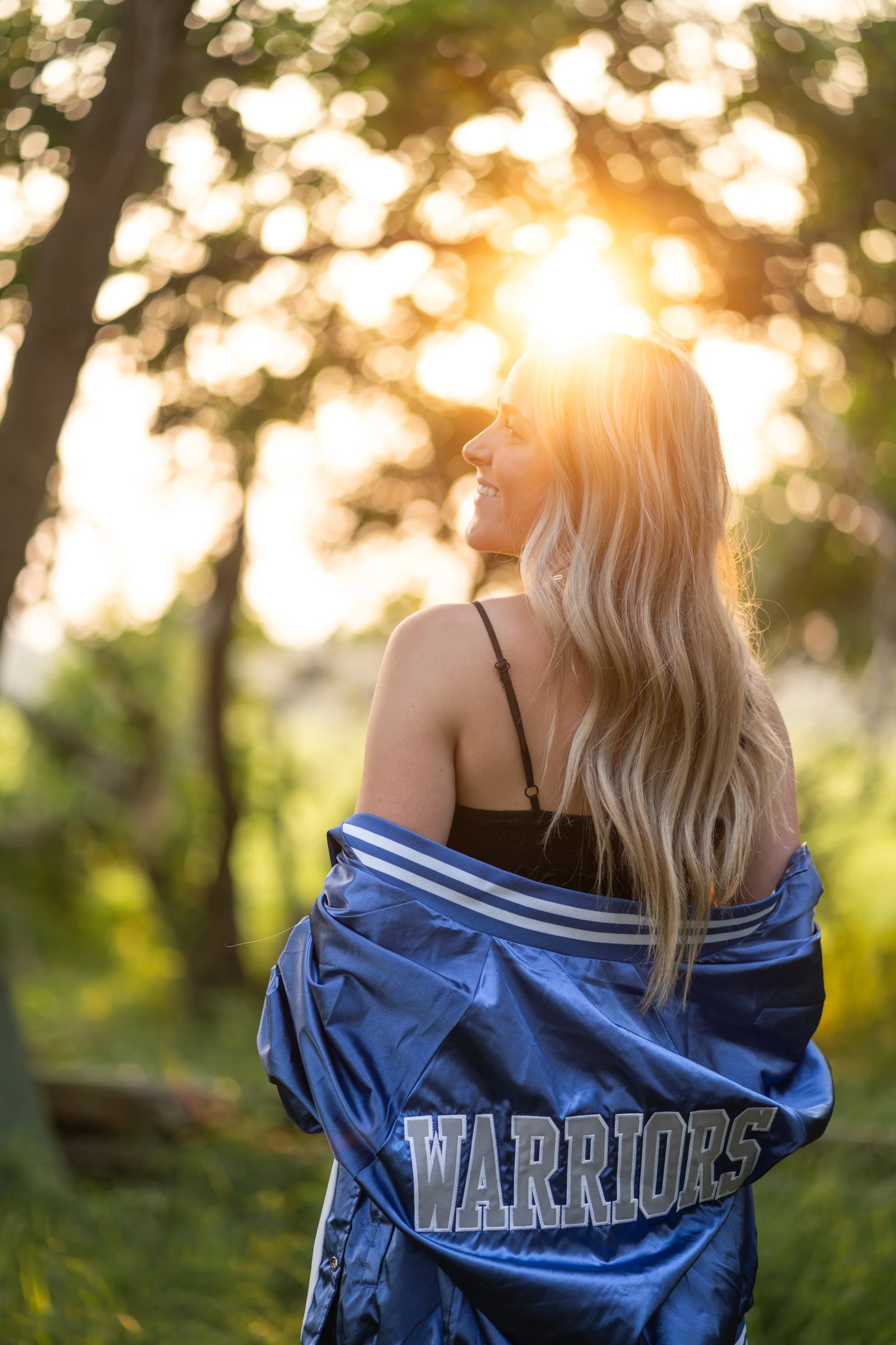 A young woman with long blonde hair smiling outdoors during sunset, wearing a black top and a blue jacket with 'WARRIORS' written on it, standing against a background of trees and bokeh sunlight.