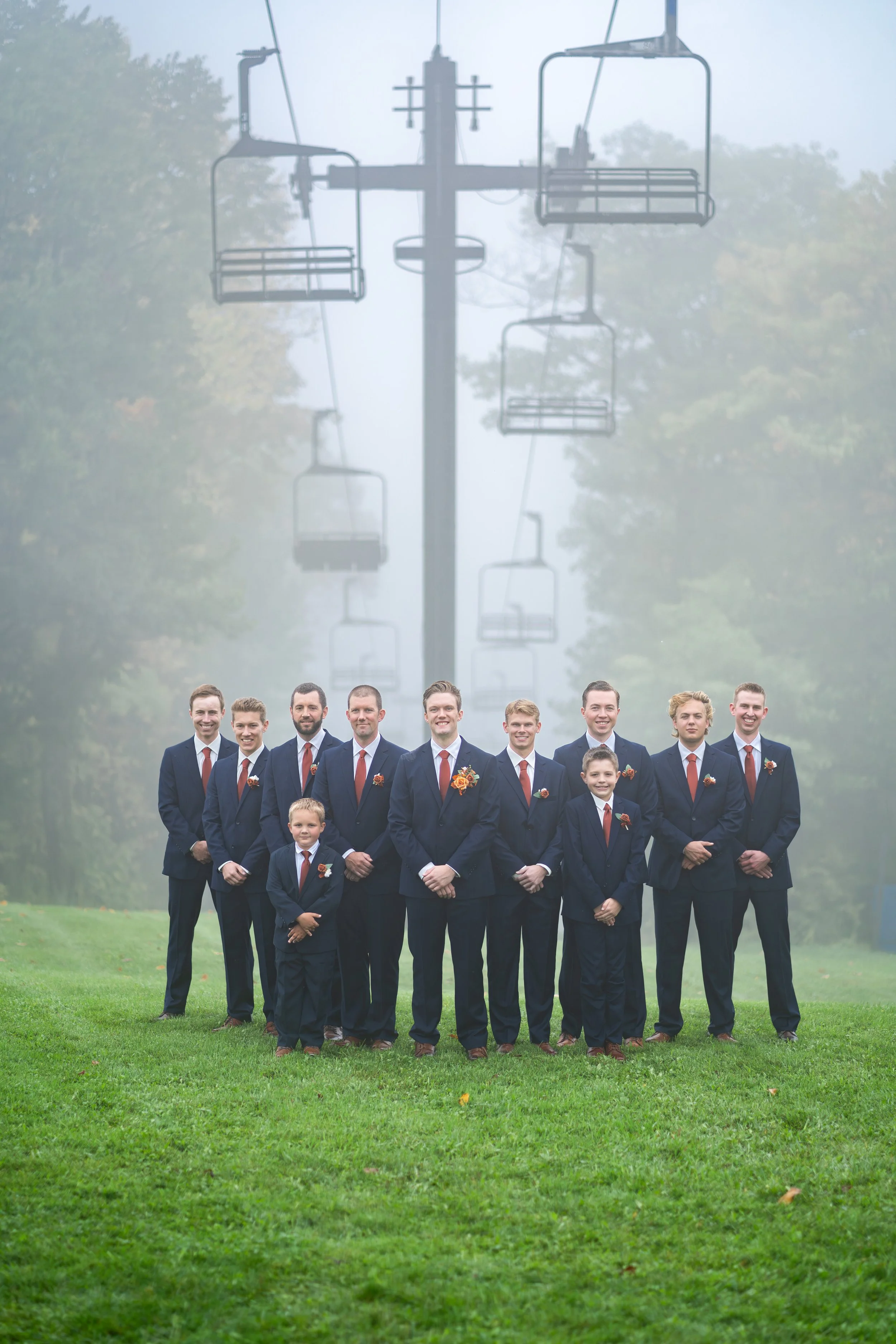 Group of men and boys wearing formal suits, standing outdoors on a green lawn, with a foggy background of trees and ski lift chairs.