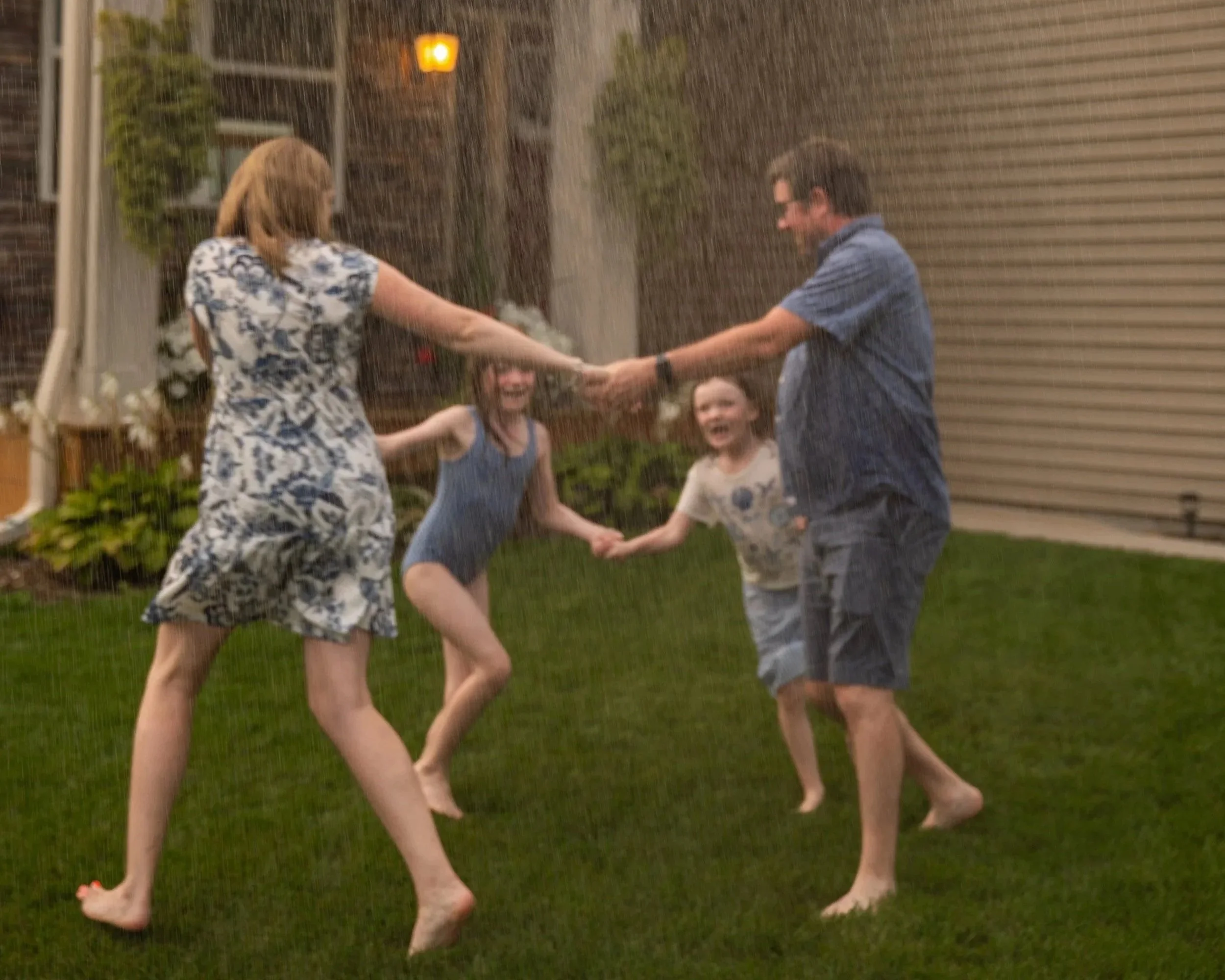 A family holding hands in a circle outdoors during a rainstorm in the evening. The parents are smiling while playing with their children on a grassy lawn near a house with siding. The rain appears heavy, creating a blurred effect in the image.