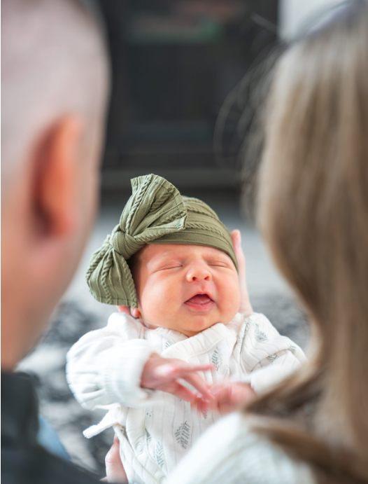 A newborn baby with a green headband, crying while being held by two adults.