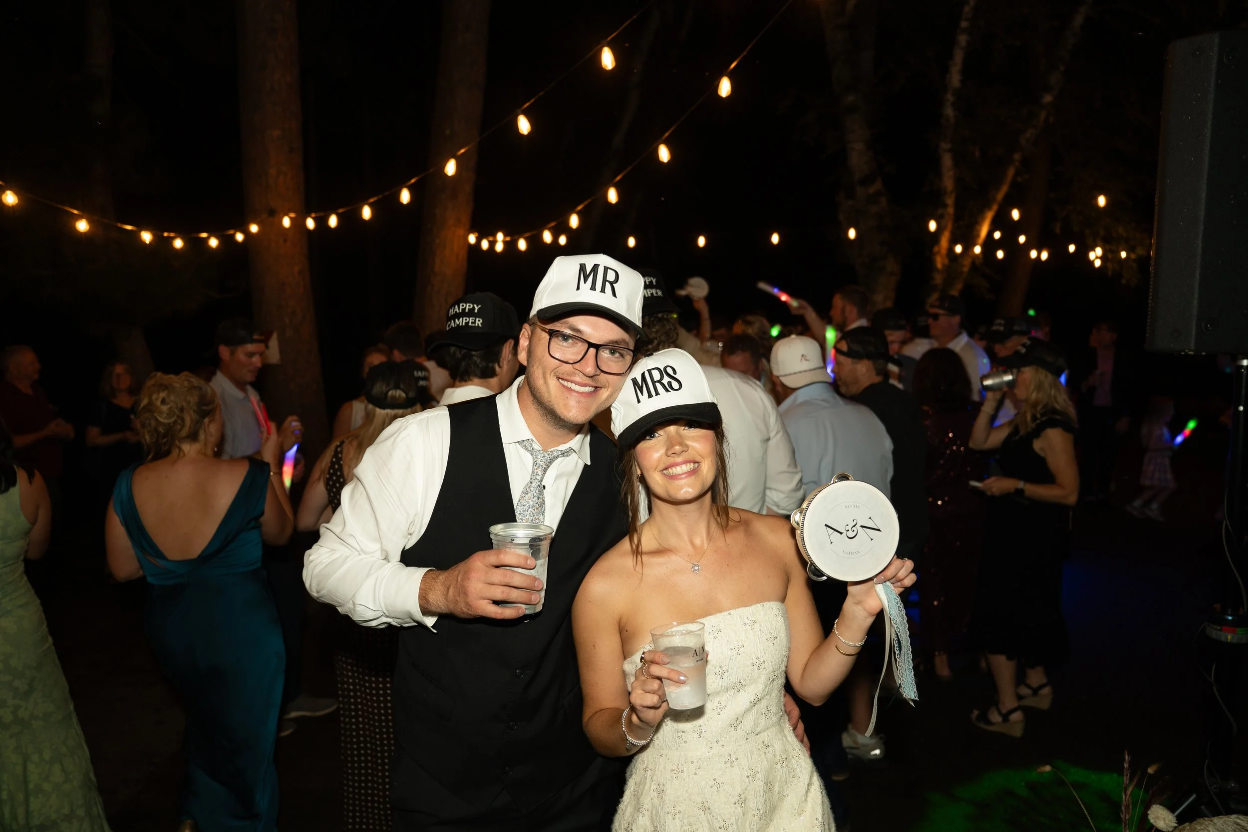 A smiling couple at a nighttime outdoor celebration, with the man wearing a "MR" hat and a suit, and the woman holding a small drum with initials and a drink, surrounded by other party guests and string lights in trees.