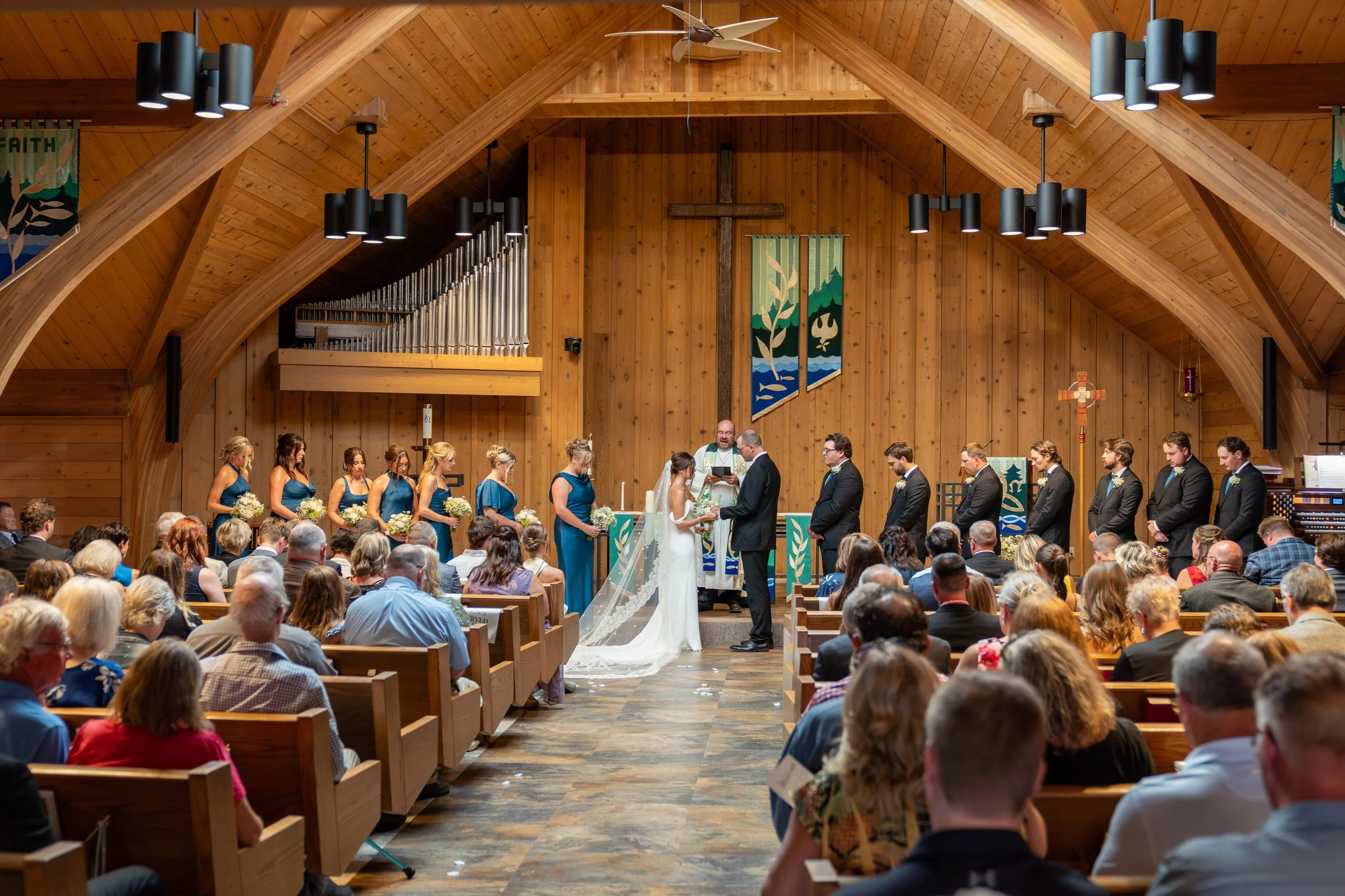 A wedding ceremony inside a wooden church with a large cross at the front. The bride and groom are exchanging vows, surrounded by bridesmaids in blue dresses and groomsmen in suits. The congregation is seated in pews, watching the ceremony.