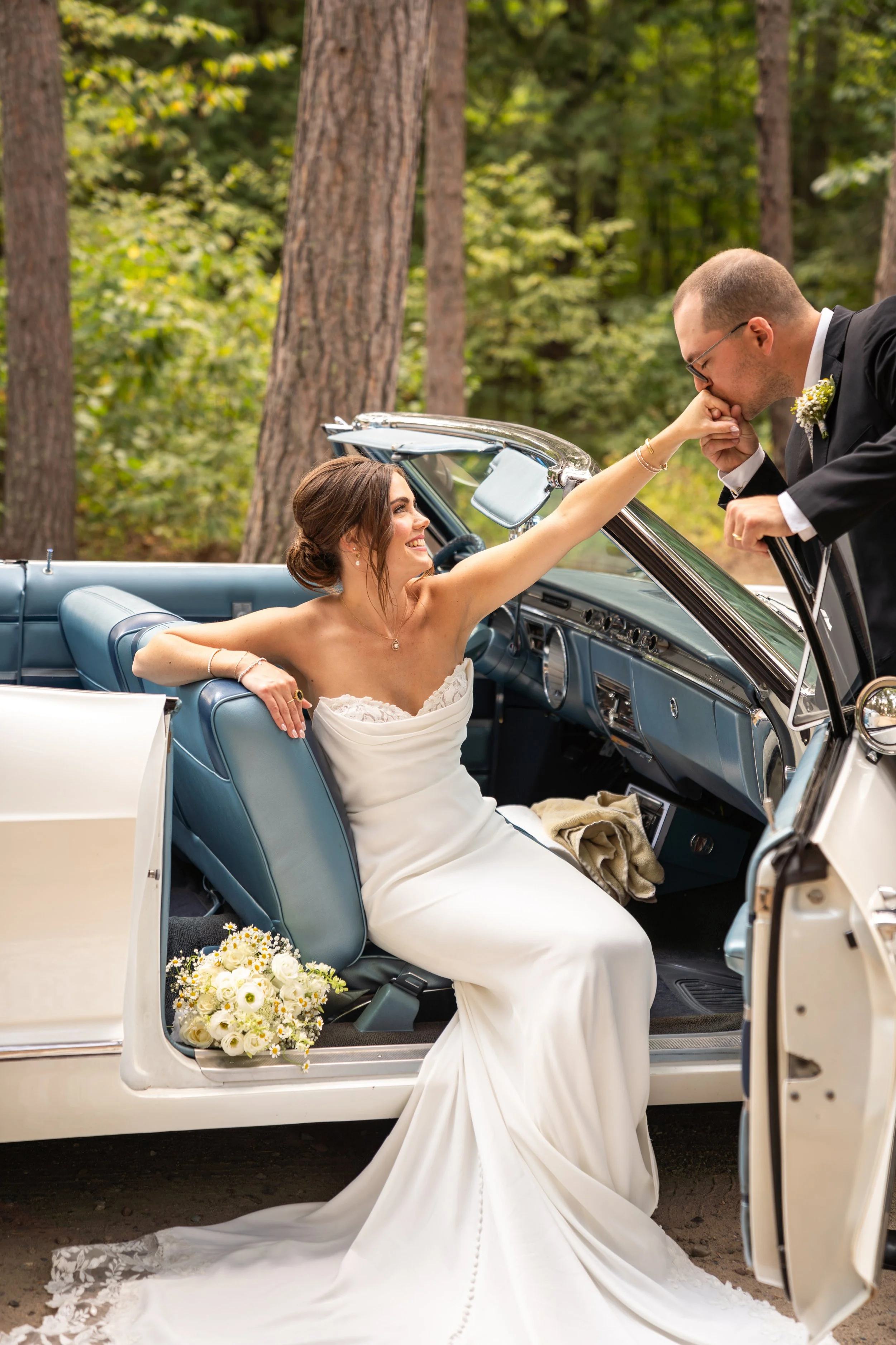 A bride in a wedding dress sitting in a vintage convertible car, smiling at her groom who is kissing her hand in a forest setting.