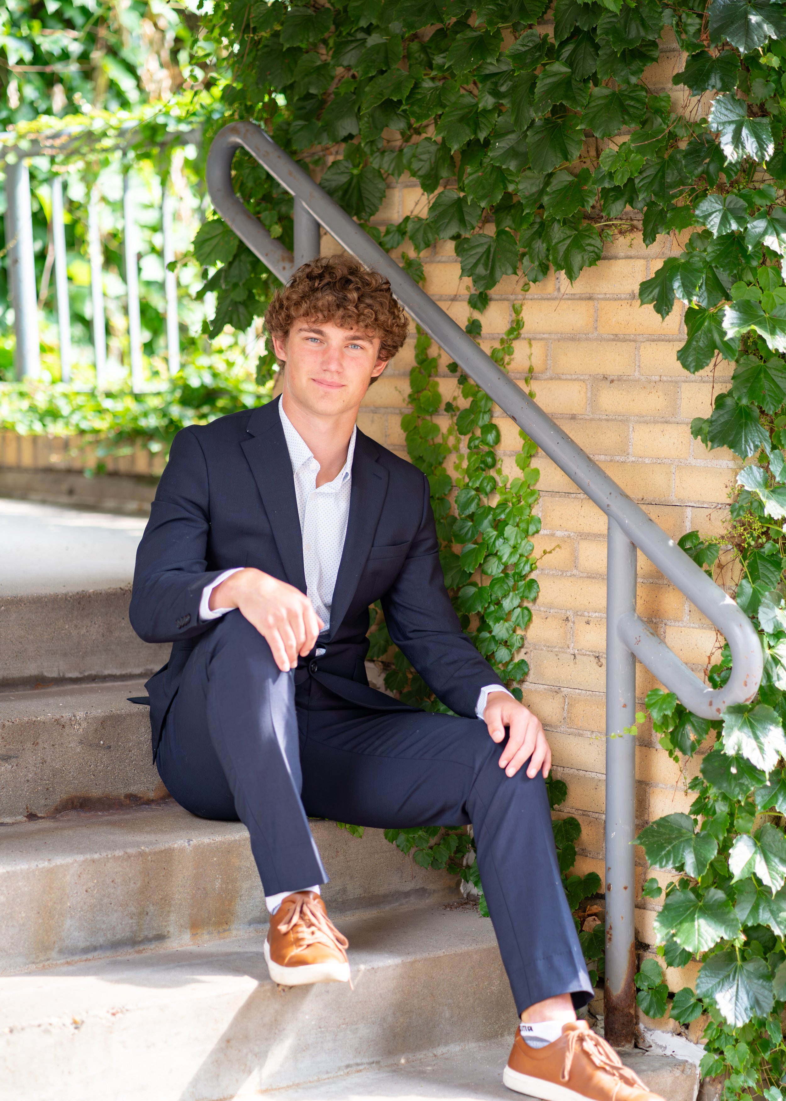 A young man with curly hair in a navy suit sitting on outdoor concrete stairs, with a green leafy vine growing on a brick wall beside him.