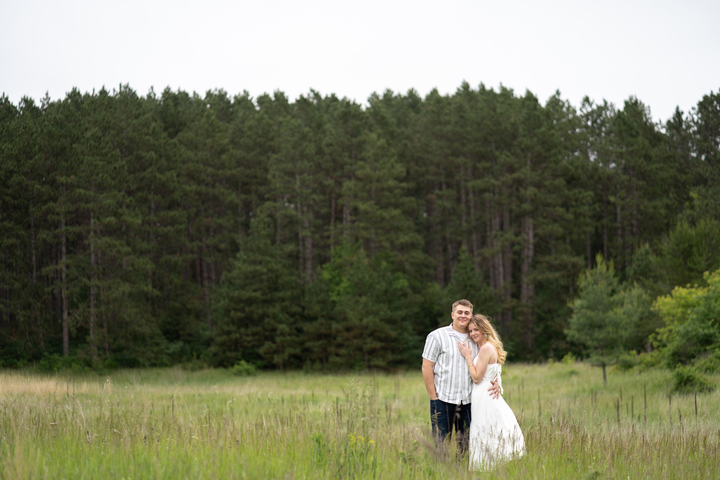 A young couple standing together in a grassy field with a large forest of pine trees behind them, smiling and embracing.