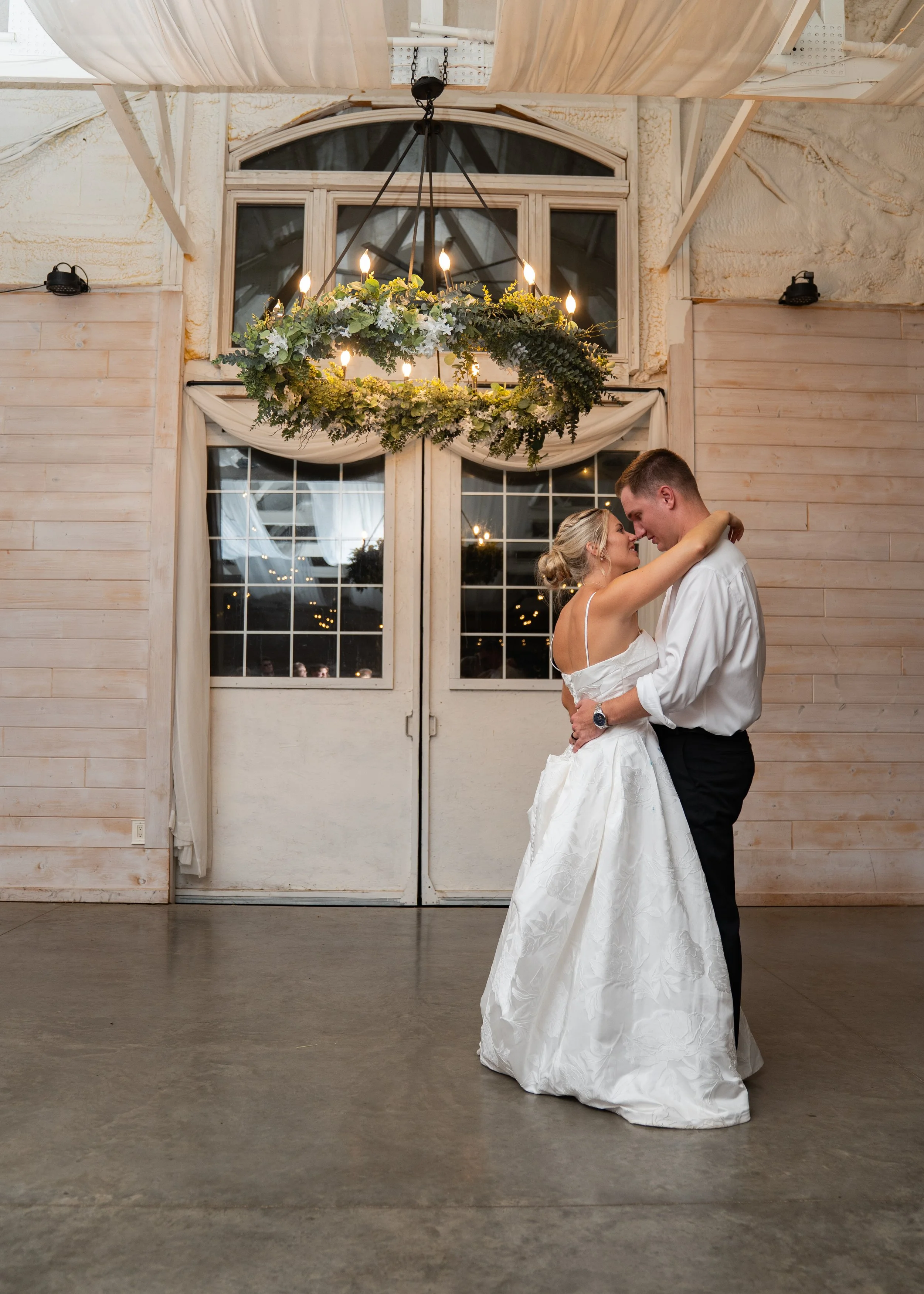 A bride and groom dancing in a venue with wooden walls, large windows, and a chandelier decorated with greenery and flowers.