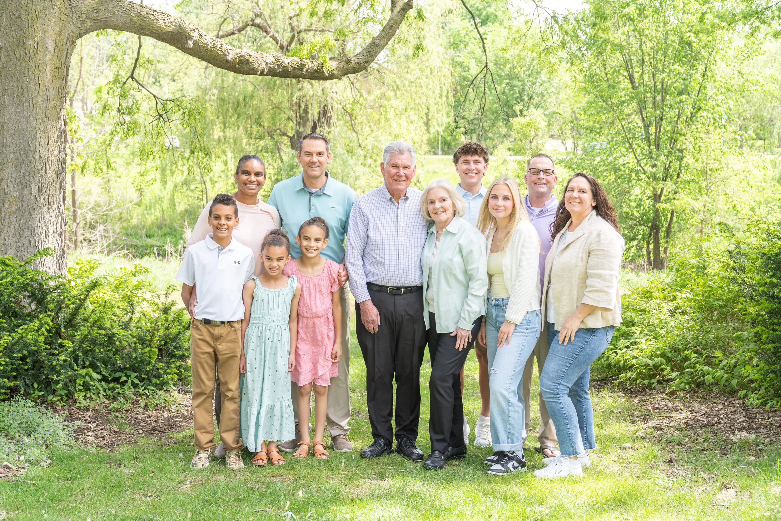 A large multigenerational family standing together outdoors in a park, smiling, with green trees and grass in the background.