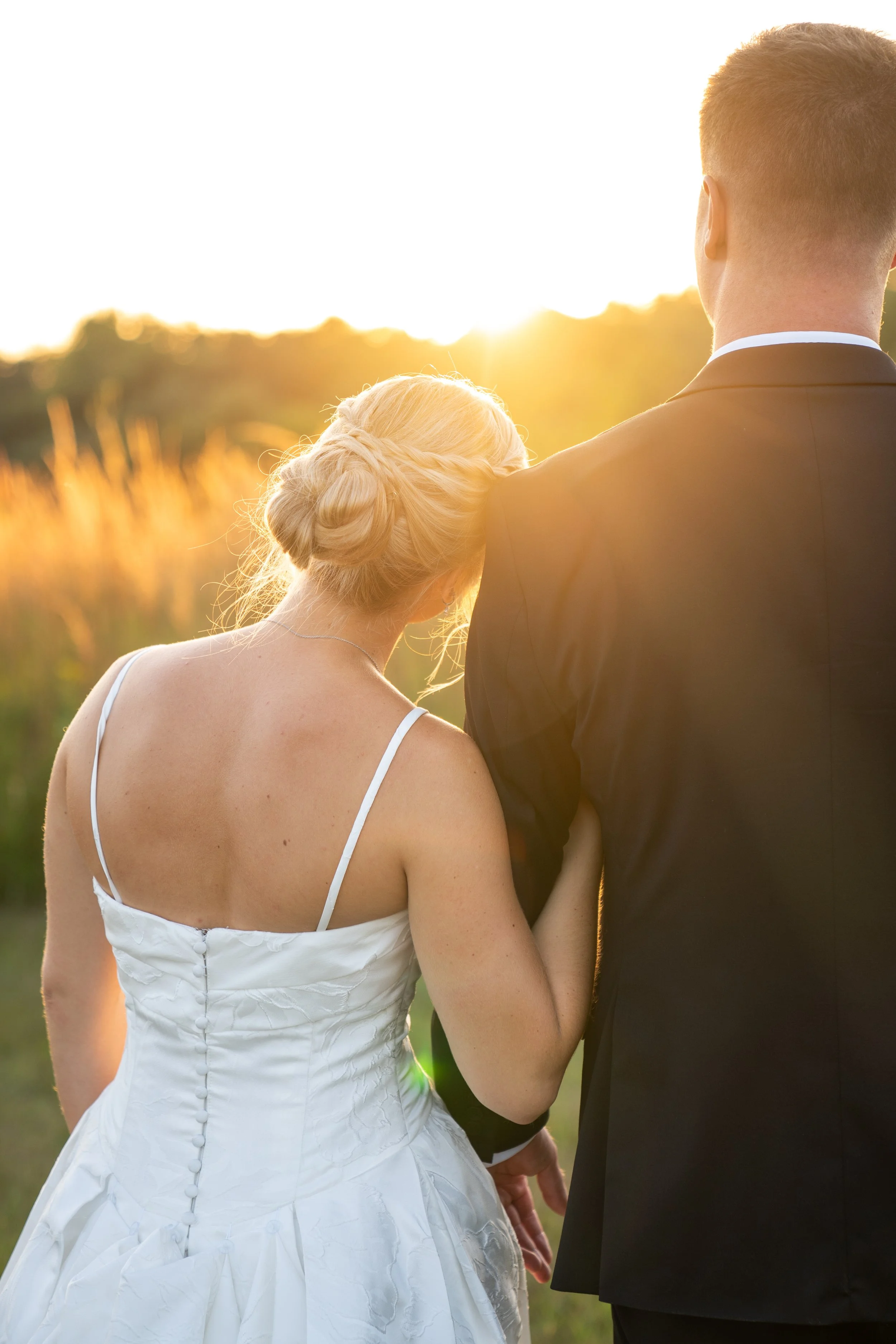 A bride and groom embrace outdoors at sunset. The bride's hair is styled in an elegant updo, and she's wearing a white wedding dress. The groom is dressed in a black suit. They are holding hands and standing close, with a bright sun setting behind them, creating a warm glow.