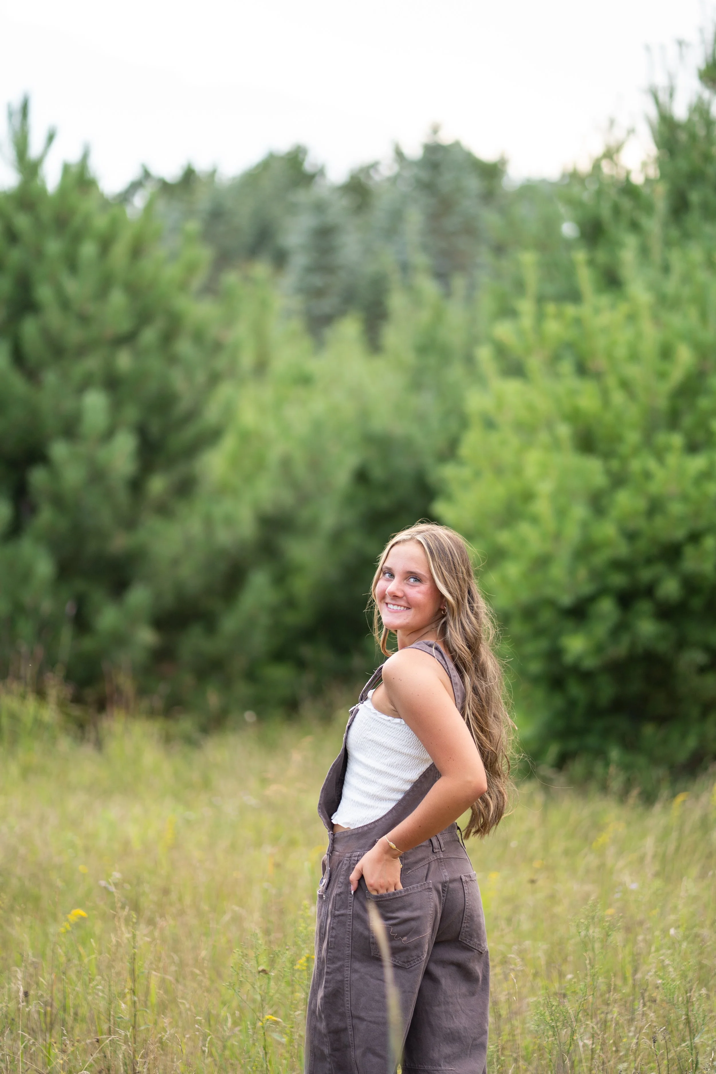 Young woman with long wavy hair wearing a white tank top and gray wide-legged pants standing in a grassy field with trees in the background, smiling and looking back over her shoulder.