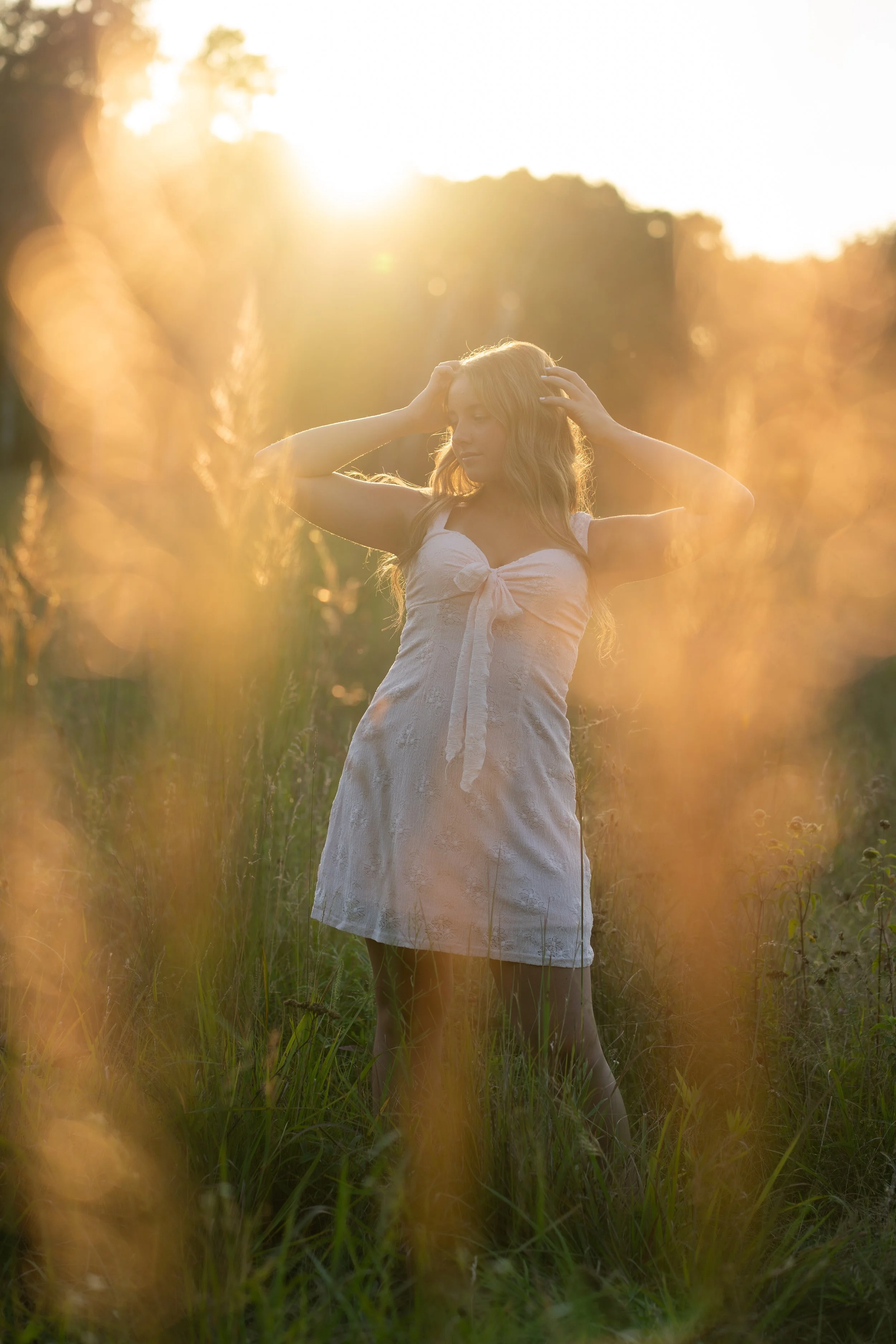 A woman with long blonde hair wearing a white dress standing in a grassy field during sunset, with her hands on her head and surrounded by tall grass and warm sunlight.