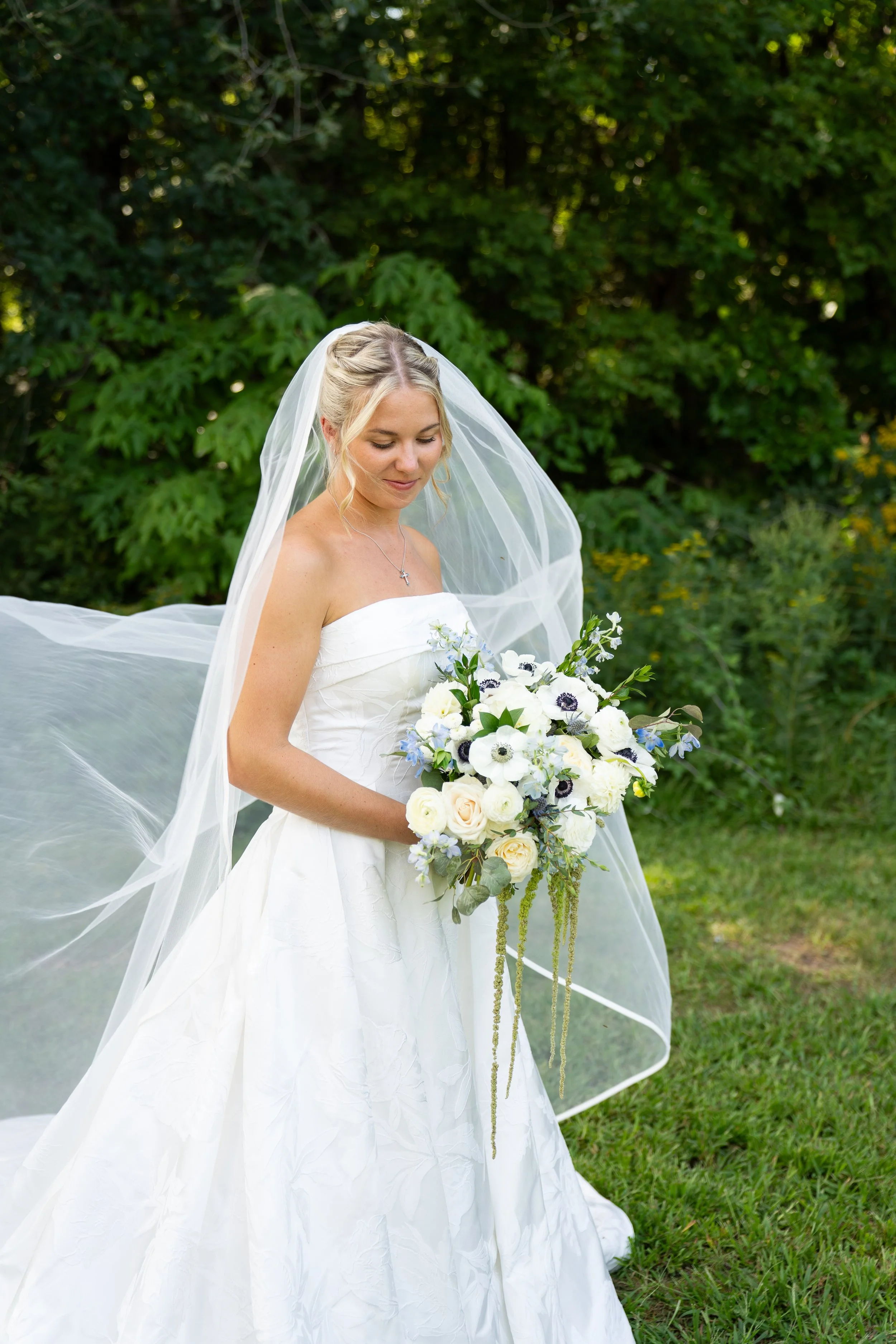 A bride in a strapless white wedding gown holding a bouquet of white and blue flowers outdoors with green foliage background.