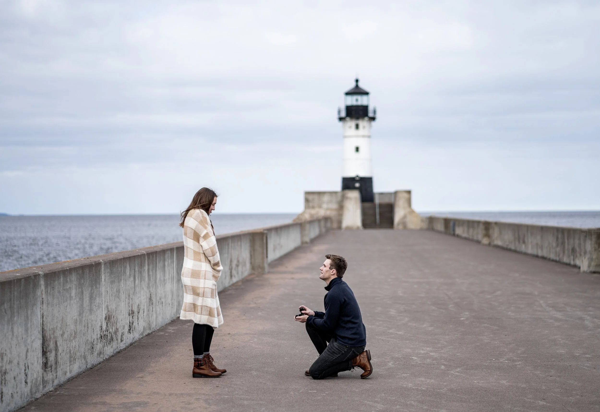 A woman stands on a pier in front of a man who is kneeling, proposing marriage, with a lighthouse and cloudy sky in the background.