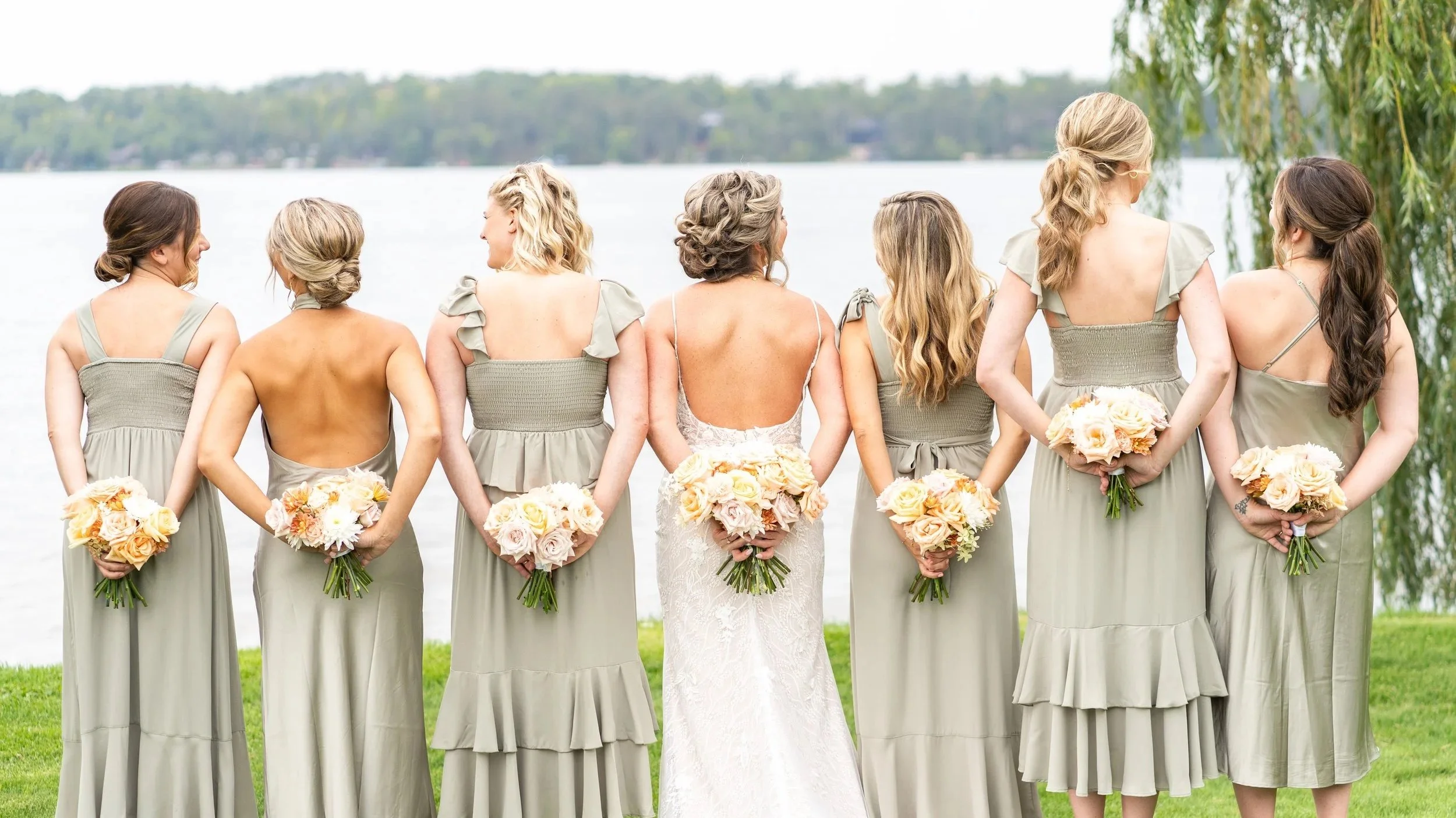A group of women, dressed in light sage green bridesmaid dresses, standing by a lake, holding bouquets of pastel-colored roses, with a bride in a white lace wedding gown among them, facing away, during a lakeside outdoor wedding ceremony.