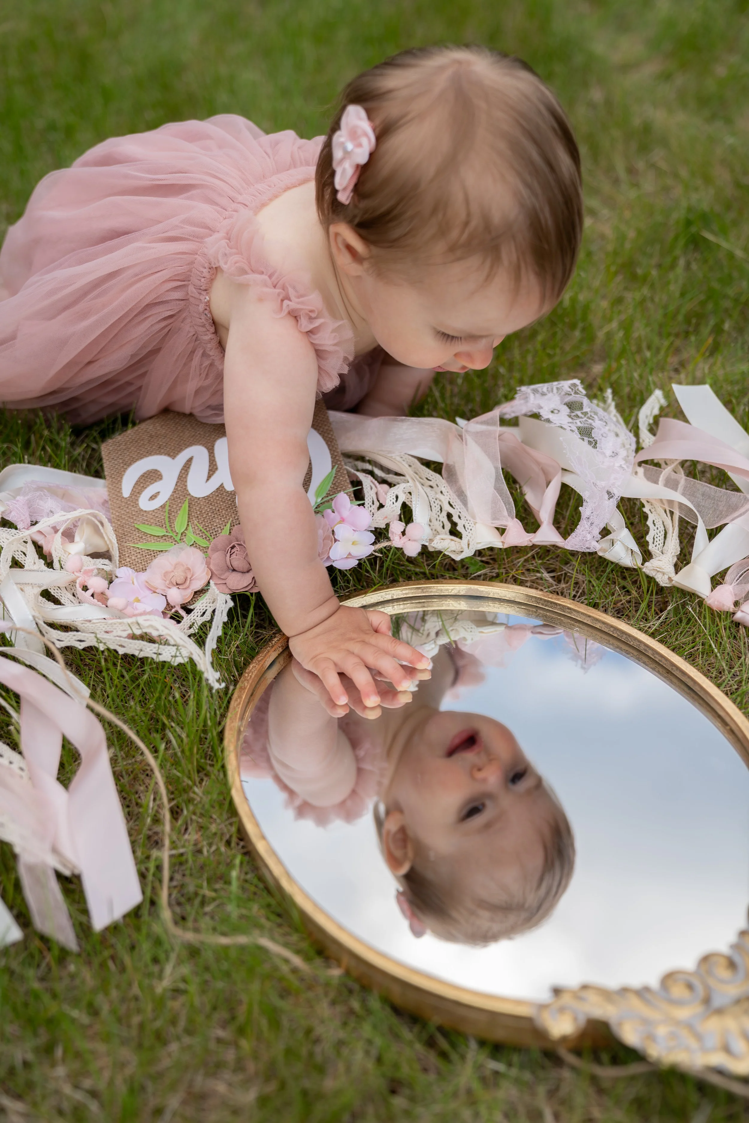 A young girl in a pink dress with ruffled sleeves and a flower hair clip is lying on the grass, looking at her reflection in a round mirror with a gold frame. She has her hand on the mirror, and there are decorative ribbons and flowers around her.