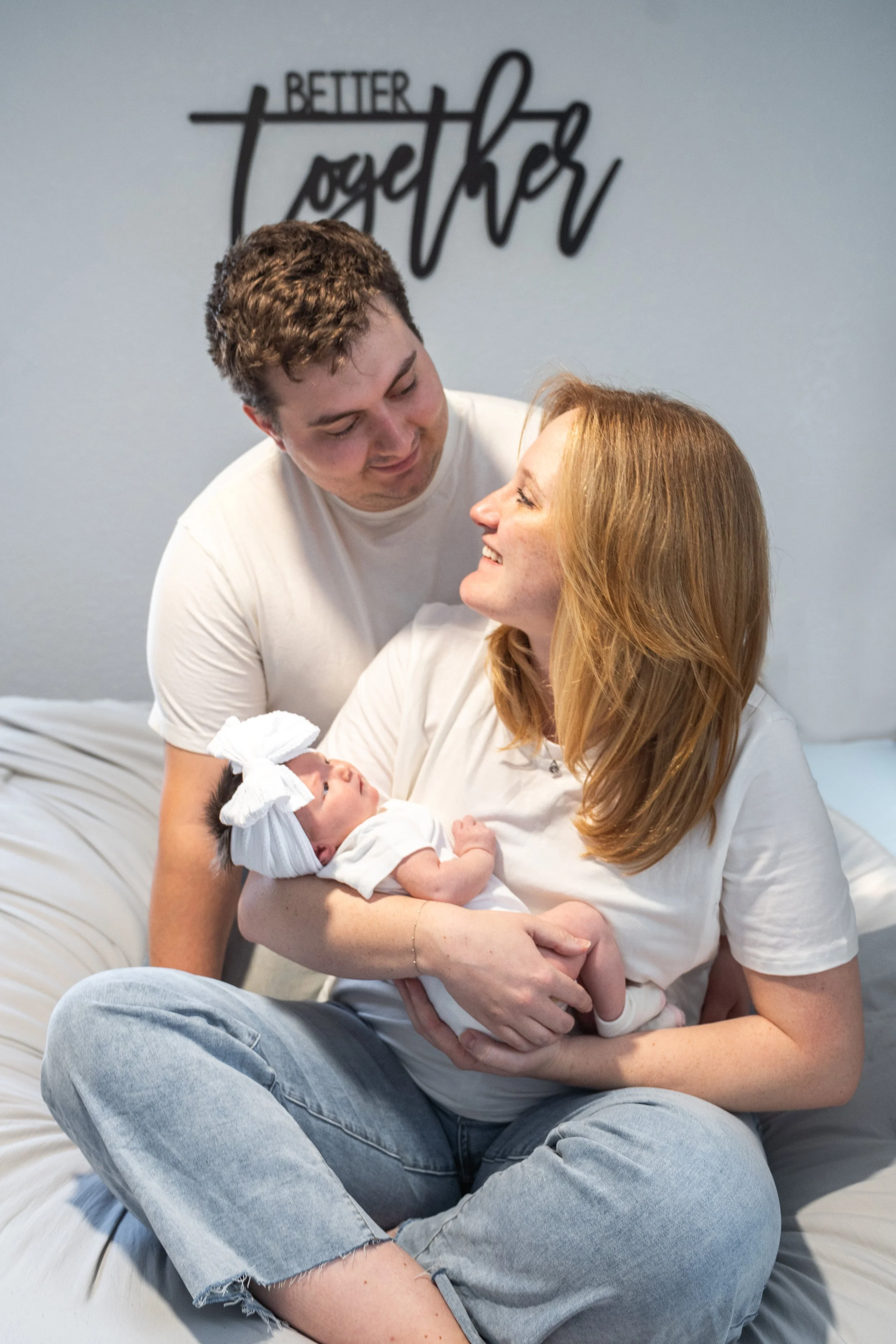 Family celebrating the arrival of a newborn baby. A woman holds the baby, while a man looks affectionately at her. The background has a sign that says 'Better Together'.