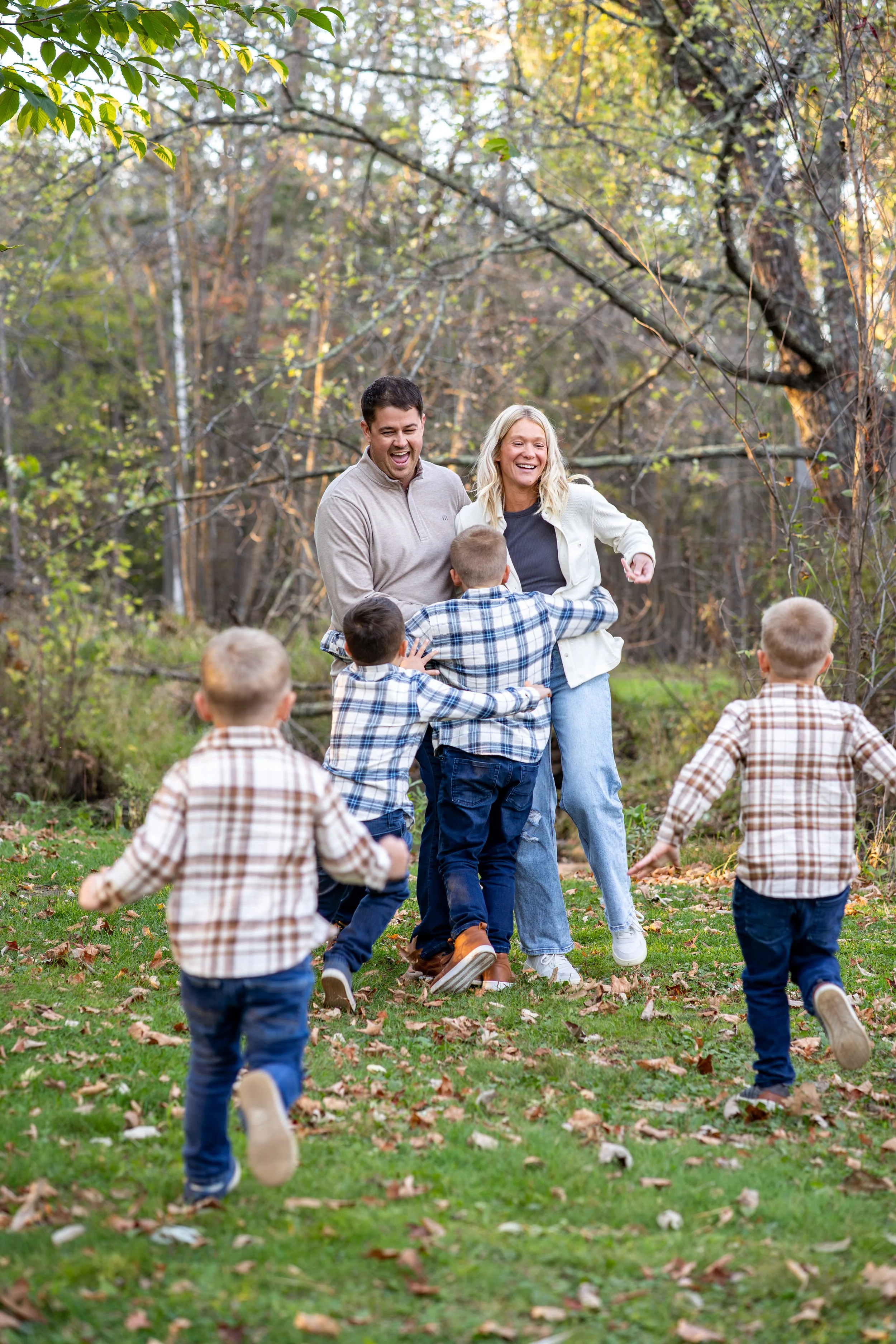 A group of children and two adults playing outdoors in a wooded area with fallen leaves during fall, smiling and enjoying a game.