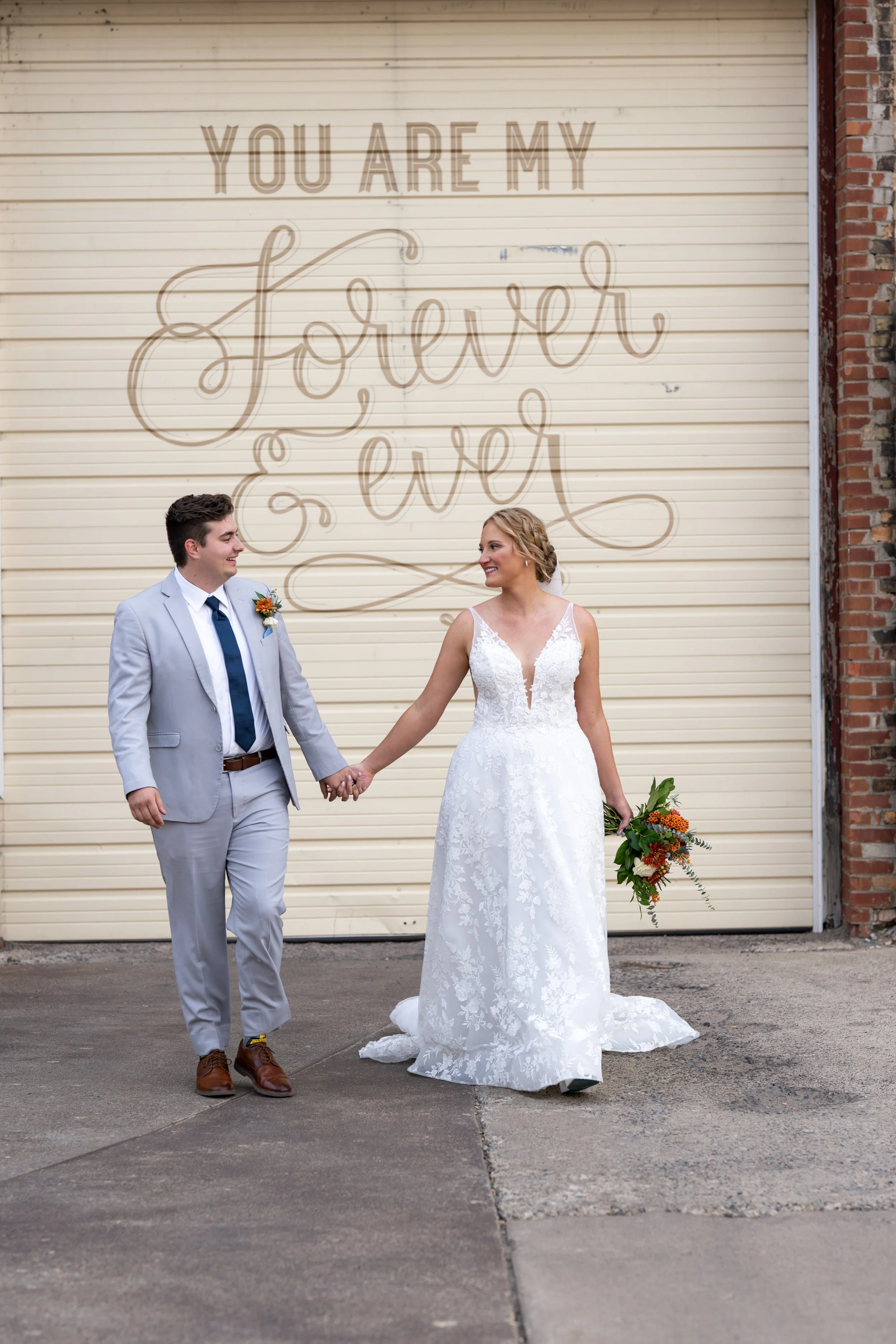 A bride and groom holding hands and smiling at each other in front of a wall with a mural that says "You are my forever". The bride wears a white lace wedding dress and the groom wears a light gray suit with a white shirt and dark tie.