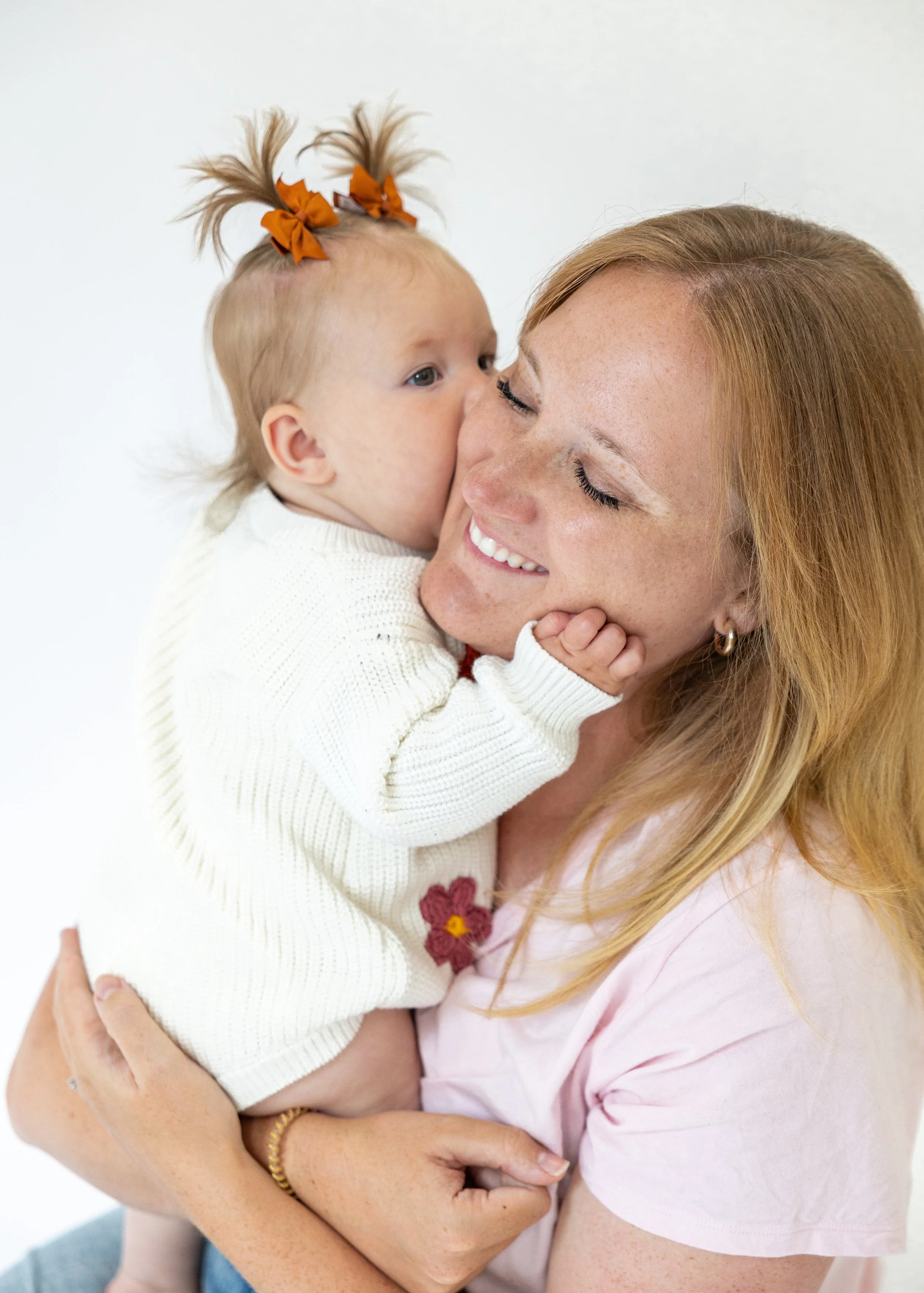 A woman with red hair smiling and holding a young girl with blonde hair styled in pigtails decorated with orange bows, while giving her a kiss on the nose against a white background.