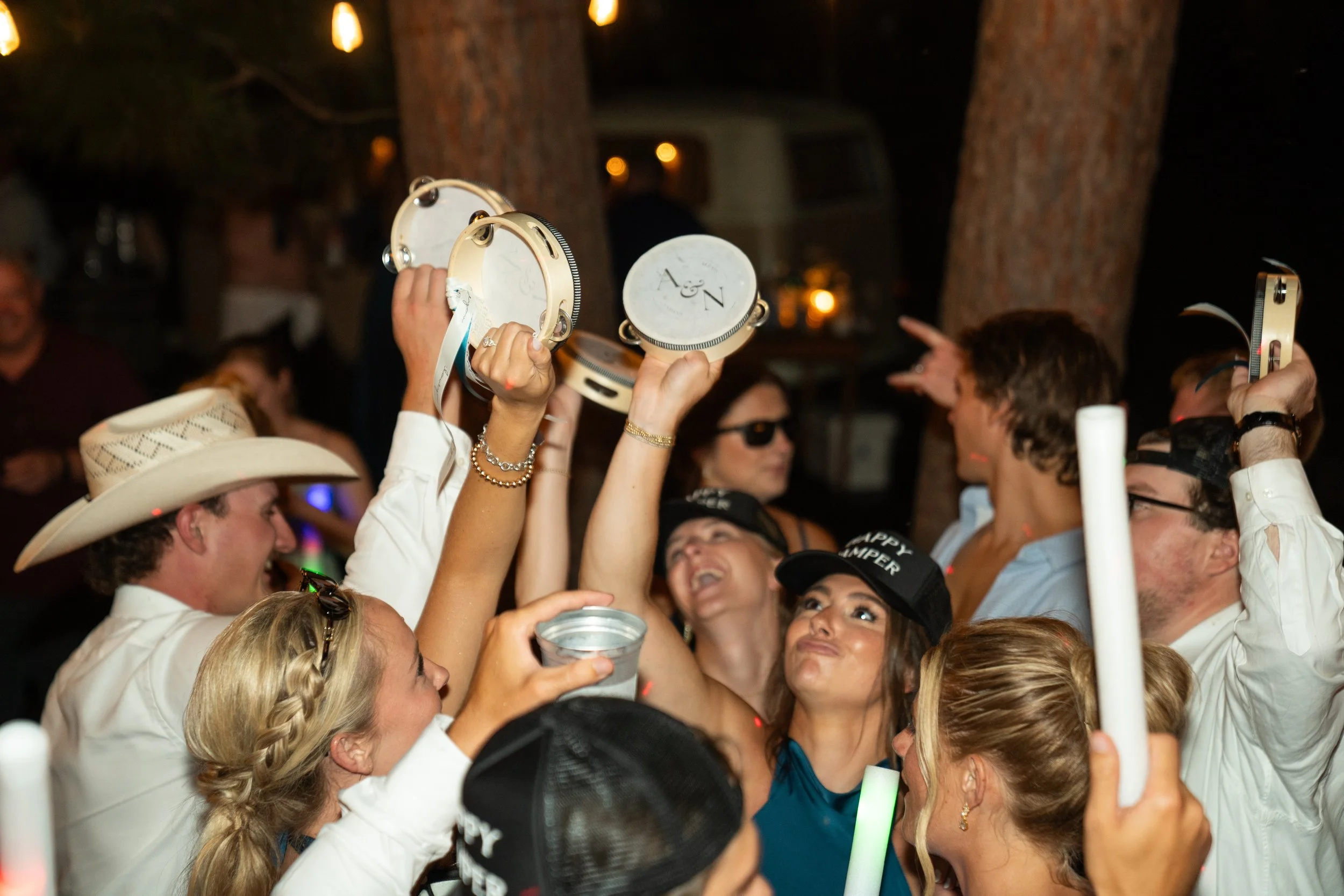 People celebrating at a party, holding small tambourines with the initials 'A & N' on them, with some wearing hats and sunglasses.