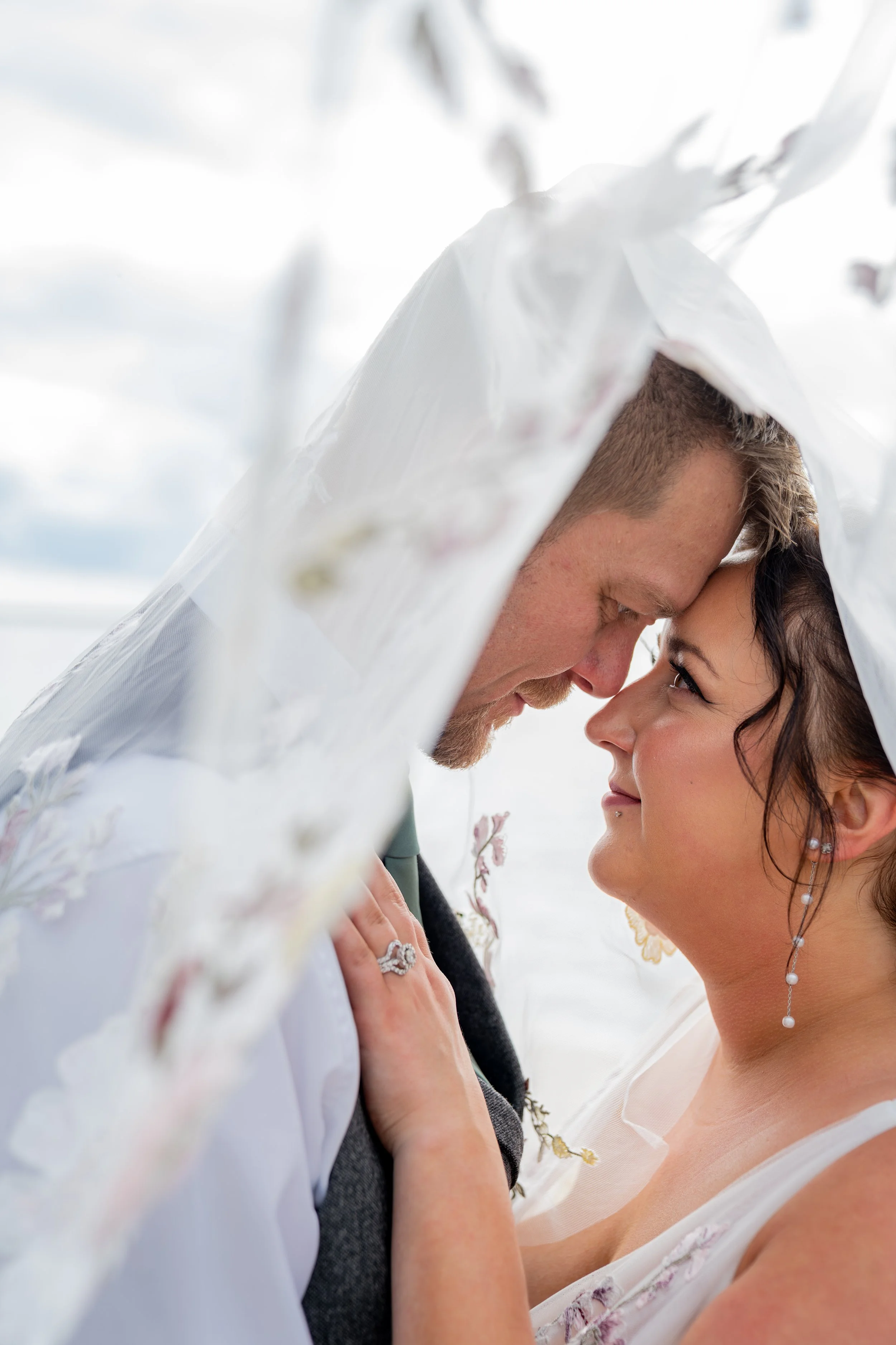 A couple face-to-face, with foreheads and noses touching, in an outdoor setting with bright sky. The woman has dark hair and wears pearl earrings, the man has short, light-brown hair. They are both dressed in wedding attire.