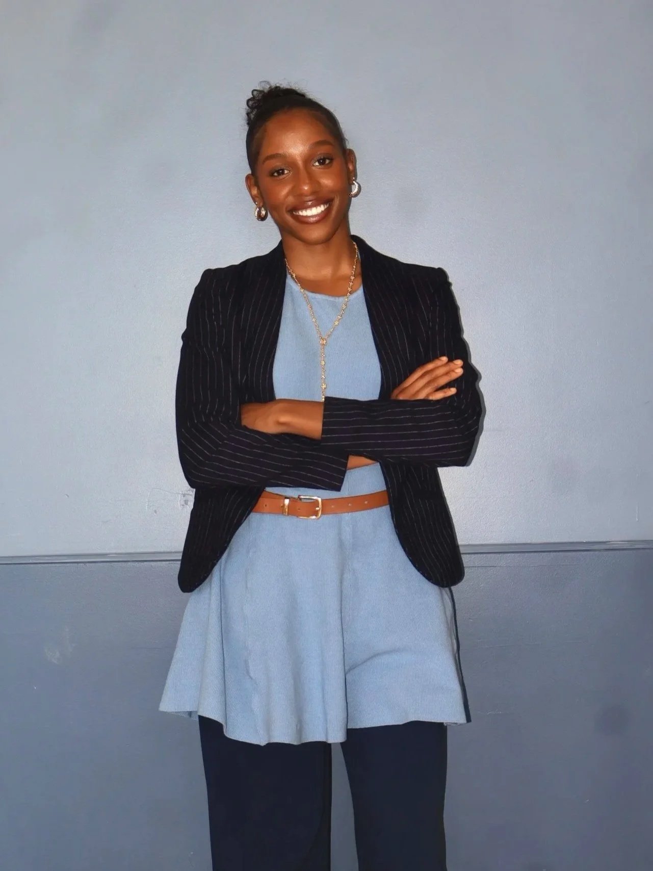 Young woman with a dark skin tone, wearing a dark pinstripe blazer over a light blue dress, crossed arms, smiling, standing against a plain gray wall.