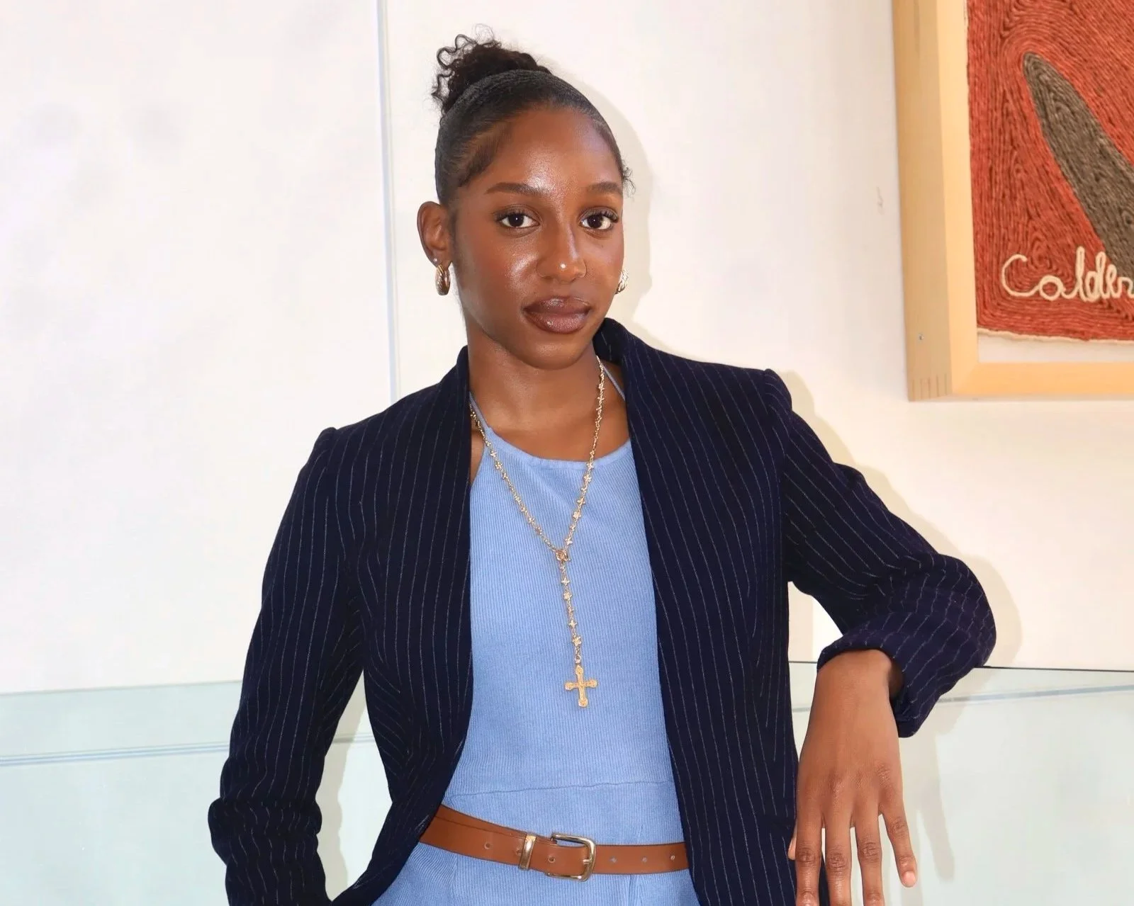 A young woman with dark skin and curly hair tied in a bun, wearing a blue dress, black pinstripe blazer, gold jewelry, and a brown belt, standing indoors against a white wall with framed art.
