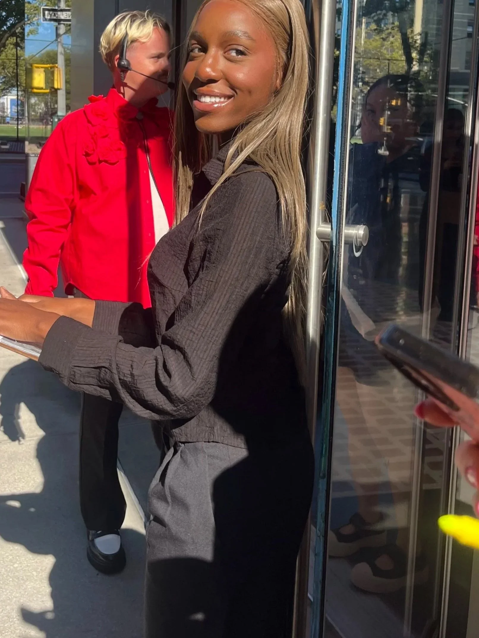 A woman with long blonde hair and a dark gray or black shirt smiling while leaning against a glass door or window. Behind her, a person with short blond hair, wearing a red jacket, a headset, and a white shirt, appears to be speaking or singing and is looking away. The scene is outdoors, with trees, a street, and a traffic light visible in the background, and the sunlight is shining brightly.