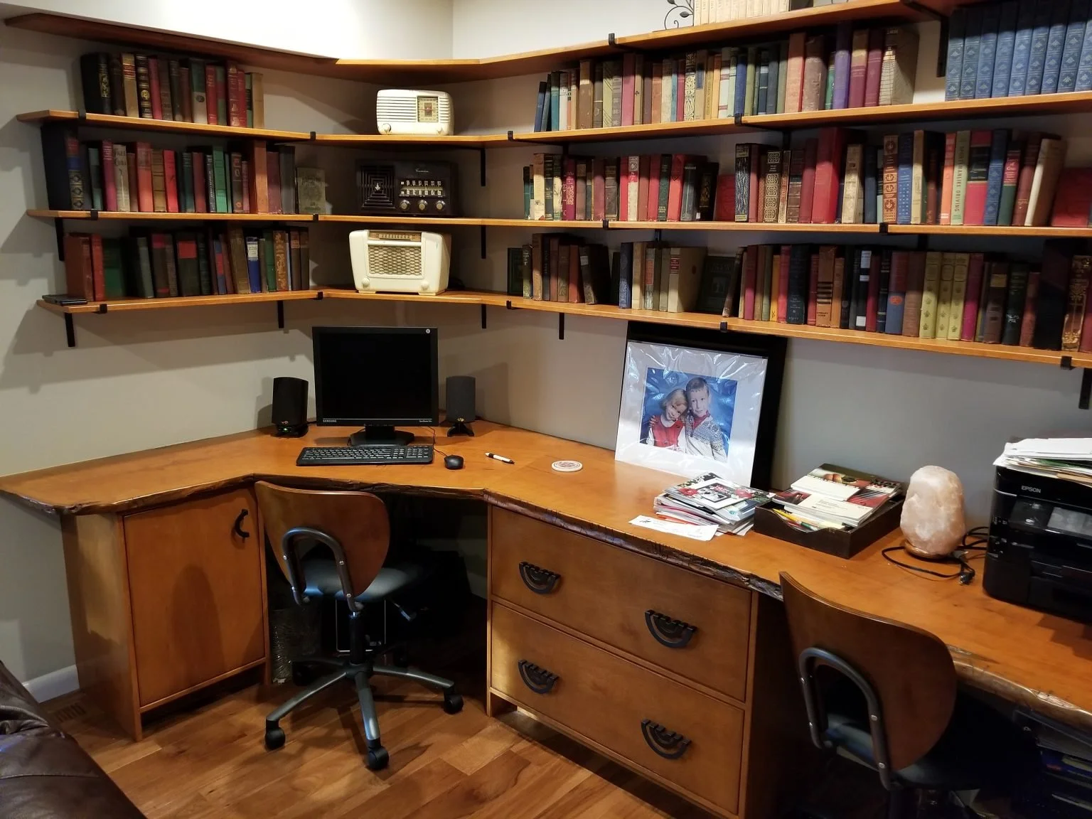 A home office corner with a large wooden desk, two chairs, a computer monitor, and two black speakers. Above the desk, there's a set of wooden shelves filled with books, a small radio, and a small white heater. On the desk, there are assorted papers, magazines, a framed photo of two children, a salt lamp, and a black printer.