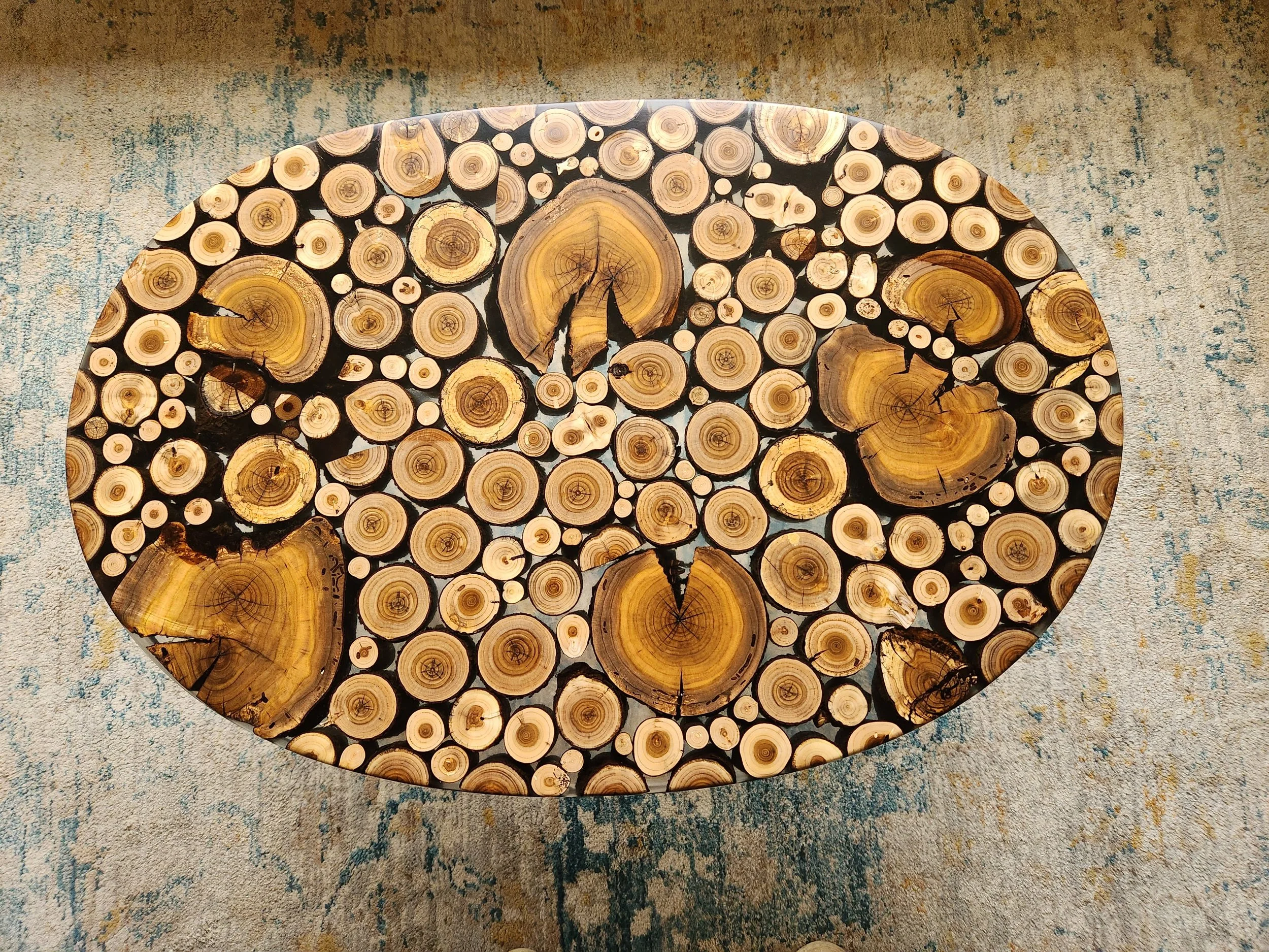 An oval custom table made of cross sections of red bud tree branches and logs in various sizes with clear epoxy, placed on a textured beige and blue patterned carpet.