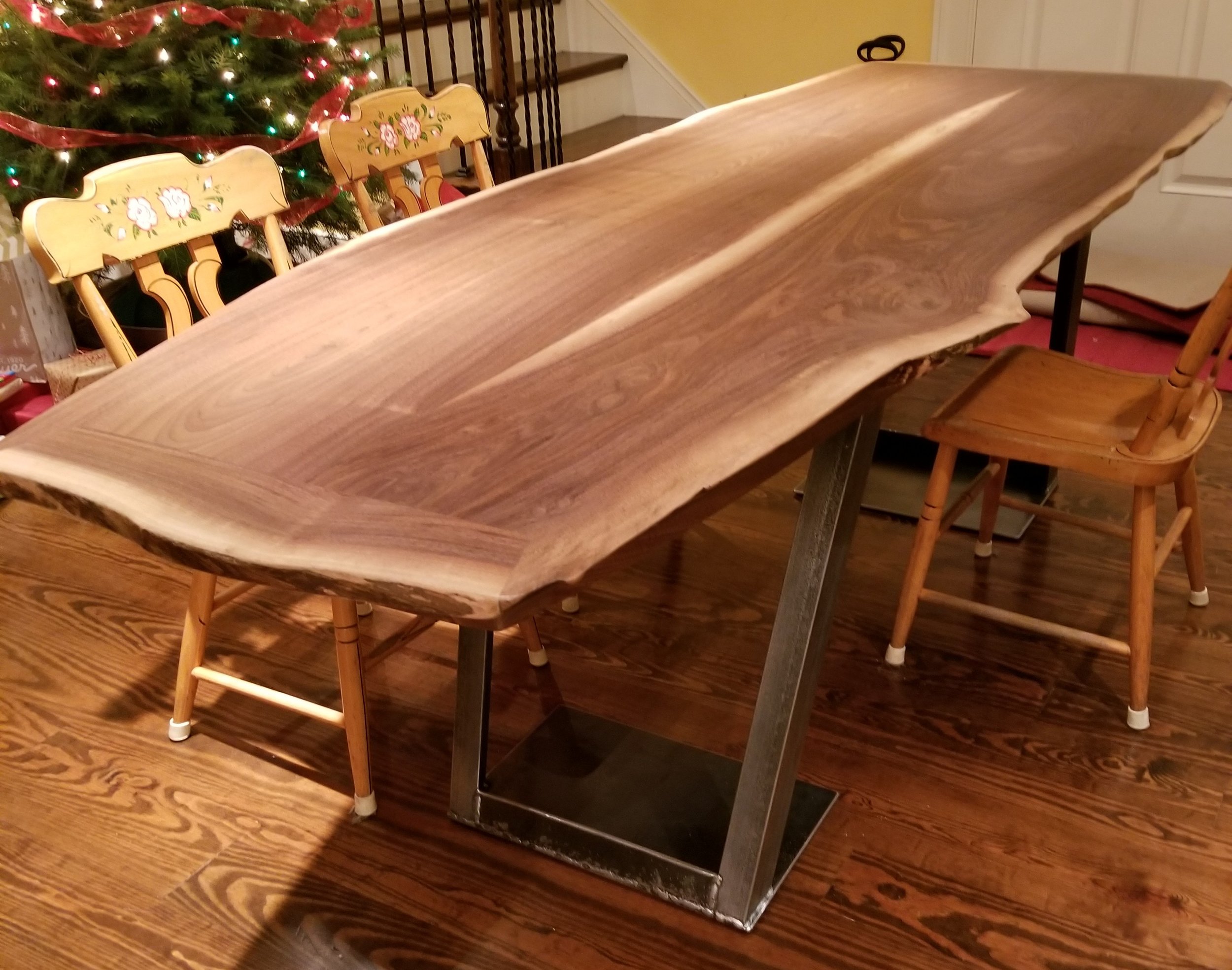 A walnut table with 2 removable leaves and a natural live edge with black metal bases in a dining room setting, with wooden chairs around it and a decorated Christmas tree with lights and ribbons in the background.